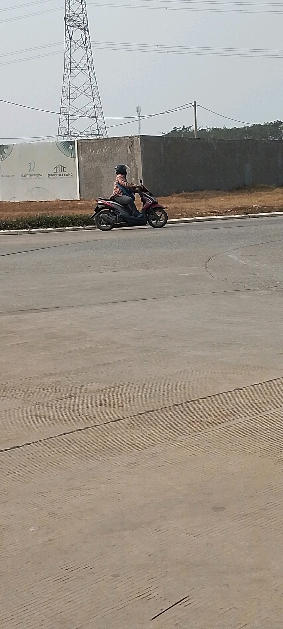 A person is riding a red scooter on a paved road. They are wearing a helmet and a plaid shirt. In the background, there is a tall tower, a concrete wall, and some trees. A billboard with text and logos is visible on the left side of the image. The environment appears to be an outdoor urban or suburban setting, possibly a construction or development area, given the billboard and wall. The lighting suggests it is daytime.
