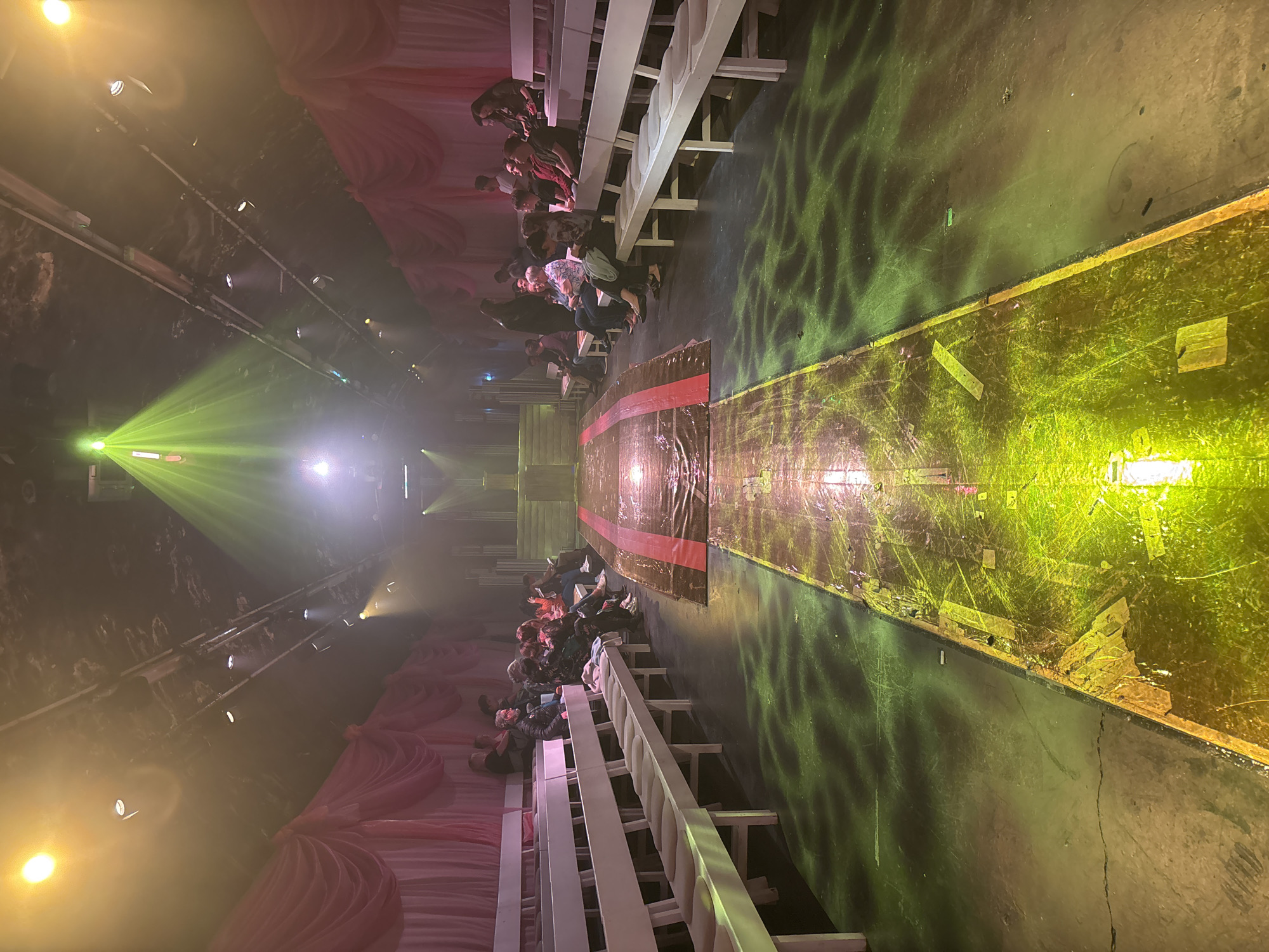 This media file depicts an indoor event, likely a fashion show or performance, situated in London, United Kingdom. The scene is characterized by a long, narrow central pathway or runway flanked by audience seating on both sides, all under dramatic theatrical lighting.
The central runway is a dark, possibly elevated, surface, appearing worn with visible scuff marks and debris. A distinct red stripe runs across its width towards the center. The runway is intensely illuminated by bright yellow and green lights, which cast dynamic patterns and reflections across its surface and the adjacent floor.
On either side of this central path are rows of white, minimalist, bench-style seating. Numerous audience members are seated here, facing inwards towards the runway, their attention directed towards the event unfolding. Their figures are discernible, though individual features are largely indistinct due to distance and the low-light conditions of the surrounding environment.
Above the seating and extending through the space, the ceiling is equipped with an array of spotlights and stage lights. These fixtures emit strong beams of green and yellow light, piercing through a visible atmospheric haze or fog, enhancing the theatrical ambiance. Sections of the walls are adorned with gathered pink or reddish fabric drapery, adding a decorative element to the otherwise dark and possibly industrial backdrop.
The artificial lighting and specific setup strongly suggest an organized public event, likely taking place in the evening or night. The overall atmosphere is dynamic and immersive. No visible text is discernible in the image.