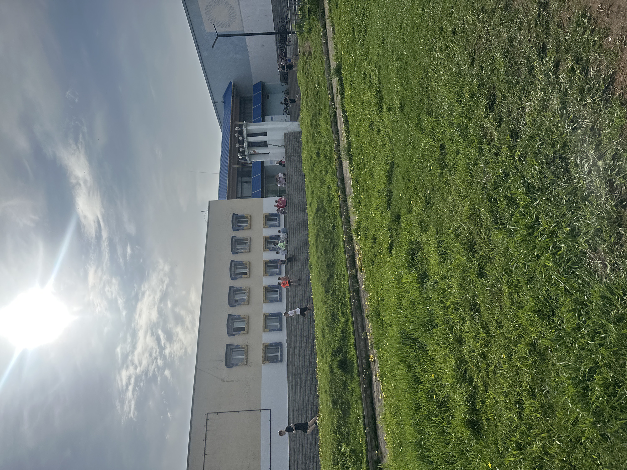 The image depicts a building next to a grassy area in Votkinsk, Russia. The building has multiple windows, some with blue trim. People are visible near the building. The grassy area appears unkempt. The sky is visible, with clouds and bright sunlight.