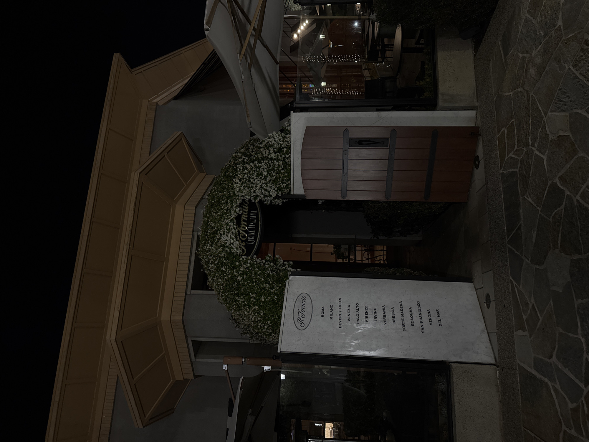 The image shows the entrance of "Il Fornaio Cucina Italiana" restaurant. The restaurant is located in Irvine, United States of America, as indicated on the sign listing city locations. The entrance is decorated with an arch of white flowers. A wooden door is to the side. A sign lists cities where the restaurant is located including Roma, Milano, Beverly Hills, Venezia, Palo Alto, Firenze, Irvine, Verbania, Brescia, Corte Madera, Bologna, San Francisco, Verona, and Del Mar. The image was taken at night. The stone walkway to the right is visible. The upper floor windows give a view of lighted interior seating. An outdoor sunshade is partially visible at the top. The building facade is tan and brown.