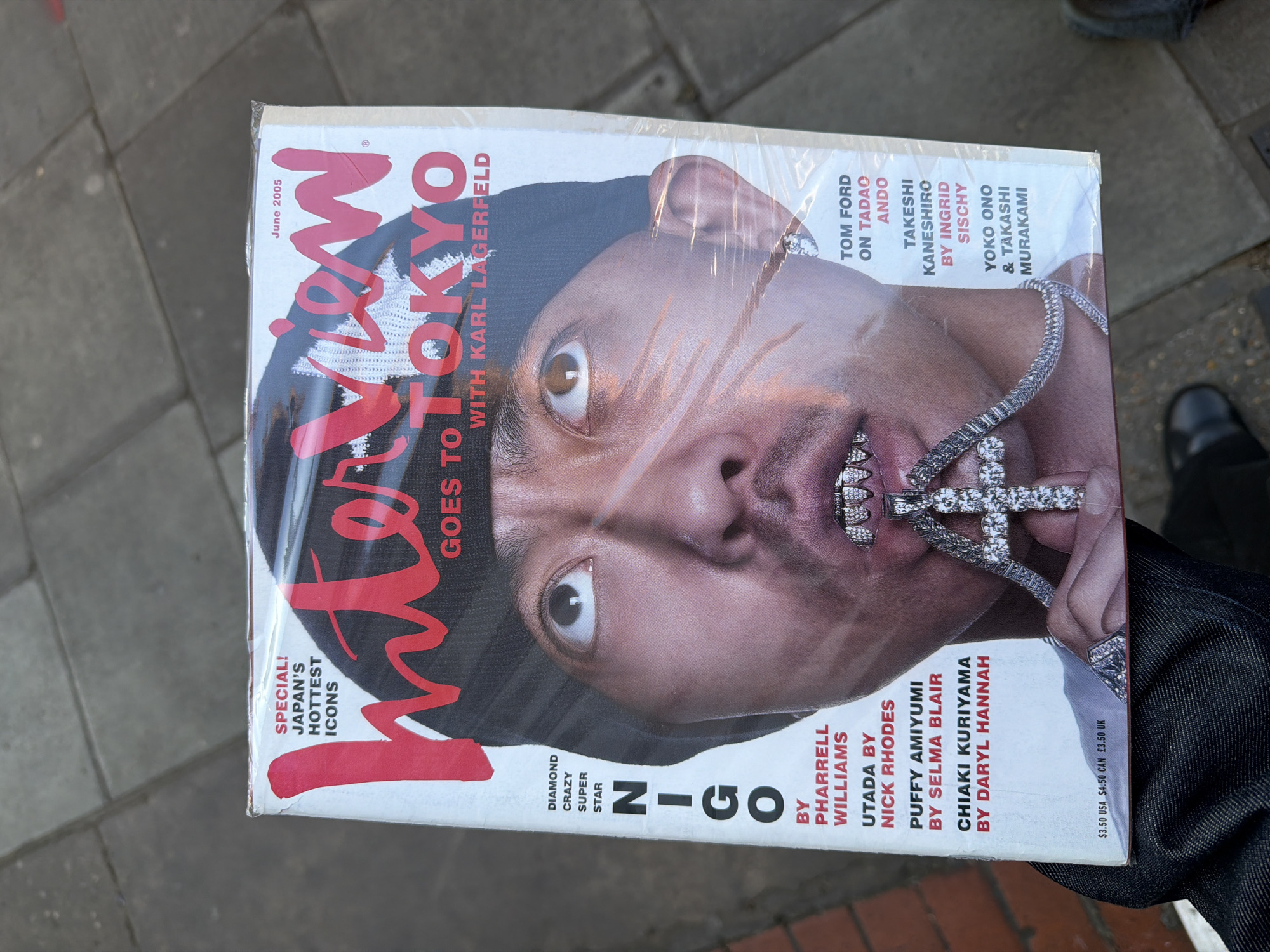 The image displays a copy of *Interview* magazine, dated June 2005, held vertically over an outdoor paved surface. The magazine is enclosed in its clear plastic wrapping, which shows some reflections and creases. The cover features a striking, close-up portrait of Nigo, looking directly at the camera with wide, intense eyes. He wears a dark knit cap, a large, intricate diamond-studded chain with a prominent cross pendant, and diamond grills on his teeth.
The magazine's title, "Interview," is in large red script. Key cover lines include "Interview GOES TO TOKYO WITH KARL LAGERFELD," and Nigo is identified as a "DIAMOND CRAZY SUPER STAR." A "SPECIAL! JAPAN'S HOTTEST ICONS" feature is also highlighted. Other listed articles and contributors include pieces by or featuring Pharrell Williams, Utada by Nick Rhodes, Puffy Amiyumi by Selma Blair, Chiaki Kuriyama by Daryl Hannah, Tom Ford on Tadao Ando, Takeshi Kaneshiro by Ingrid Sischy, and Yoko Ono & Takashi Murakami. The price is listed as "$3.50 USA $4.50 CAN £3.50 UK," confirming its international distribution.
A person's hand, part of an arm, and a leg clad in dark denim are visible holding the magazine. The background consists of grey, interlocking pavement stones, characteristic of an urban sidewalk. A dark shoe or boot is partially visible in the upper right corner of the frame. The lighting suggests natural daylight. Based on the "£3.50 UK" price and the provided context, the scene is set in London, United Kingdom.