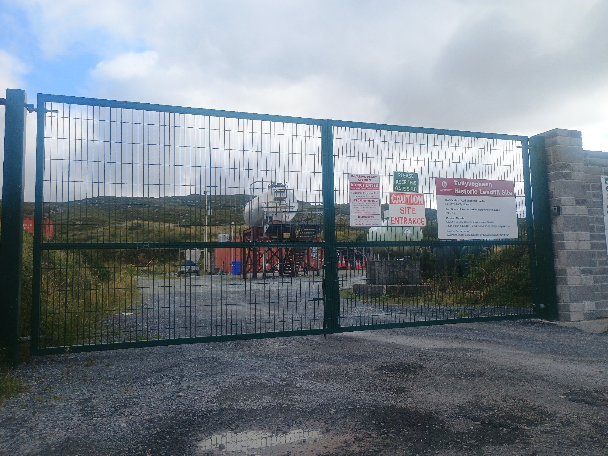 An exterior, eye-level shot captures a green metal double-door gate, partially open, that is protecting a landfill site. The gate is lined with a silver metal mesh. Mounted on the gate are several signs: a red and white sign that reads "CAUTION SITE ENTRANCE," a red and white sign that says "INVASIVE PLANT SPECIES DO NOT ENTER," and a white sign with red text reading "Tullyvoghbeen Historic Landfill Site." To the left of the gate is a tall, dark green metal post, and to the right, a short, grey brick wall extends. Beyond the gate, a gravelly path leads into the site, where two large white industrial tanks are visible. Further into the site, a green grassy hill rises under a cloudy sky.