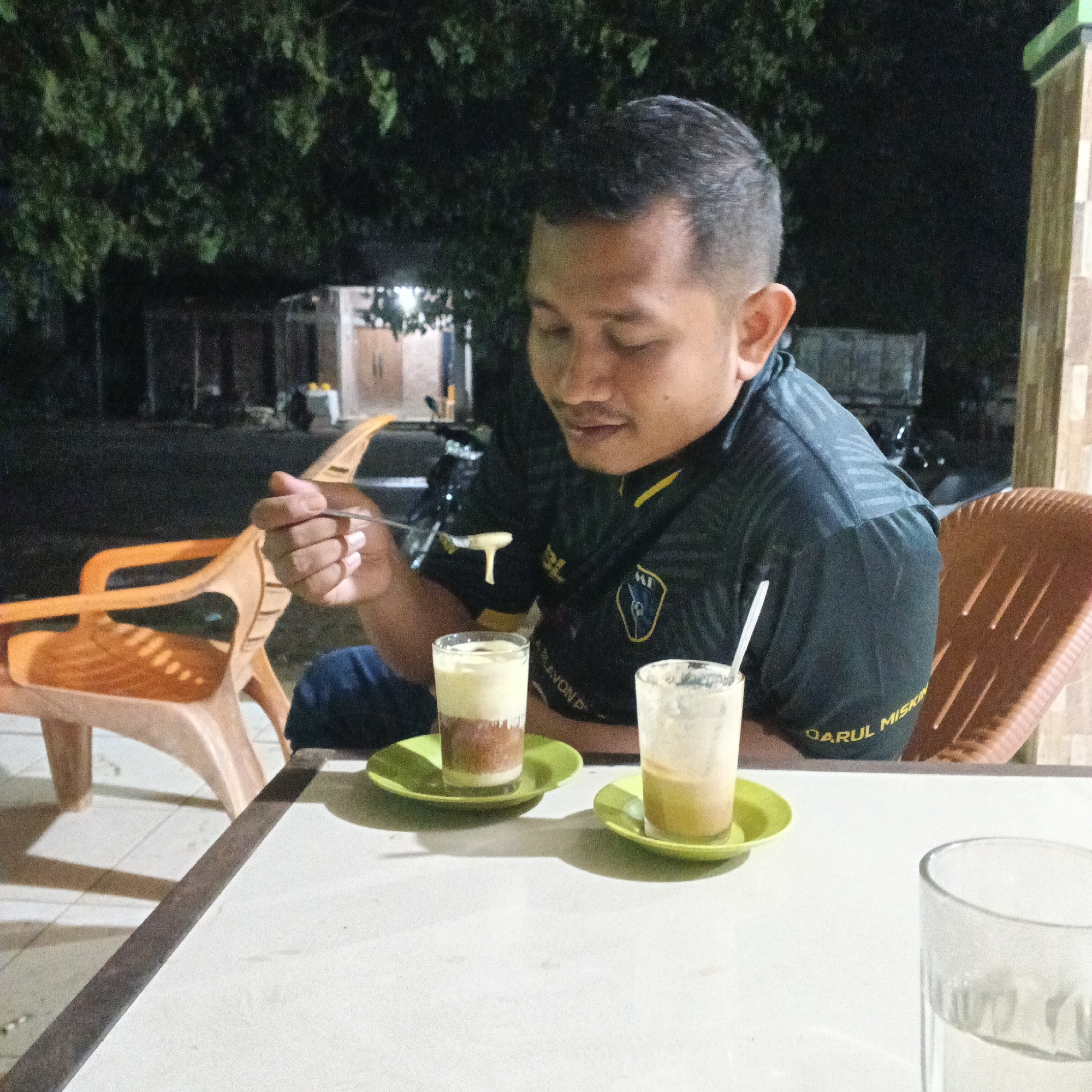 In an outdoor setting at night, a man is seated at a table with two glasses of beverages. He is wearing a dark t-shirt with yellow accents and appears to be eating something from a spoon held in his right hand. In front of him are two glasses, each containing a layered drink, placed on green saucers. One glass is fuller than the other and has a straw. The table is white and appears to be made of plastic. In the background, there are some trees and what appears to be a building with lights. An orange plastic chair is visible to the left of the man, and another brown plastic chair is to his right. The ground is tiled and some shadows are cast on it. The overall scene suggests a casual dining or social gathering outdoors.