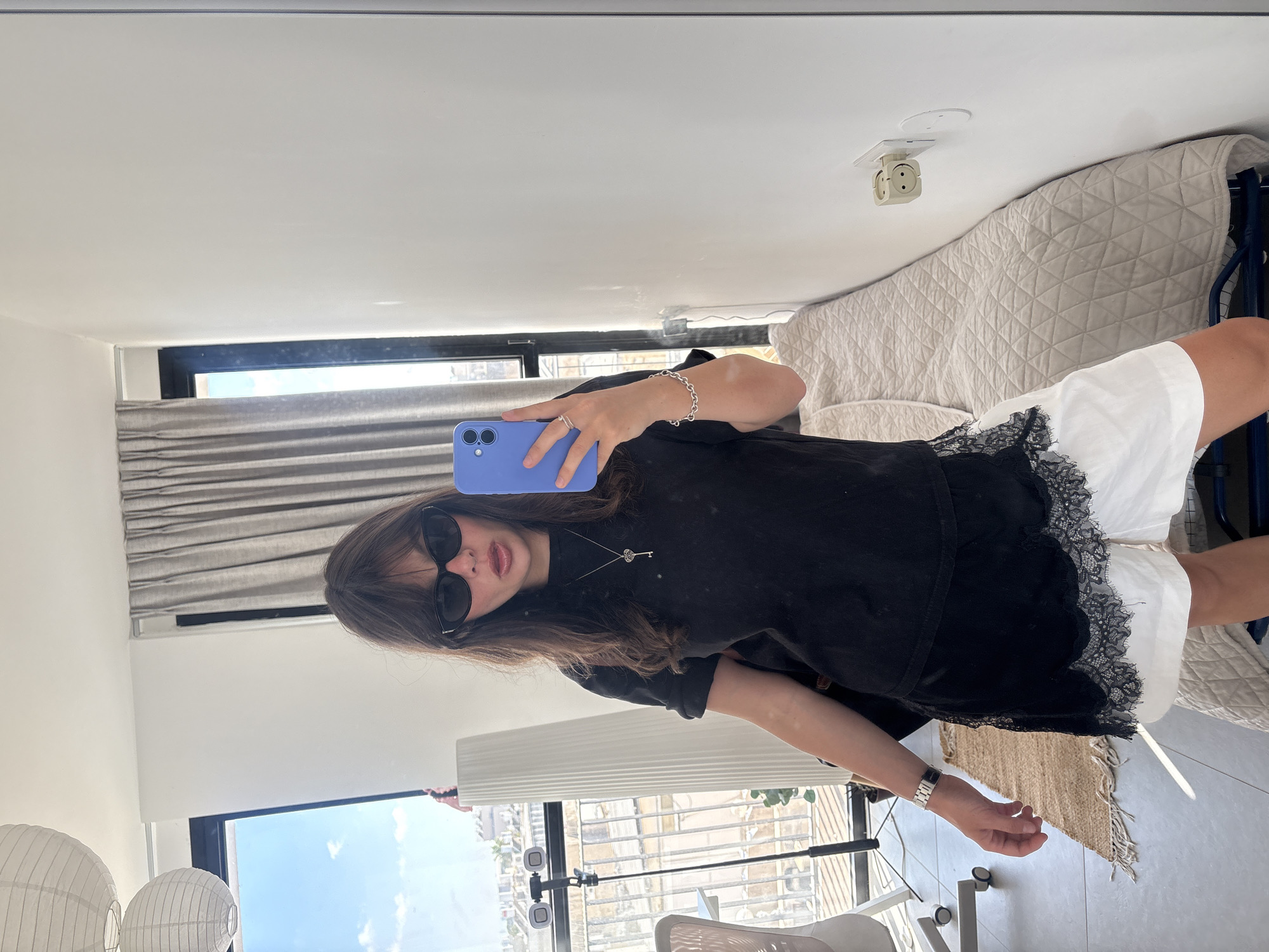 A young woman with dark hair and bangs, wearing sunglasses, a black t-shirt, and white shorts with black lace trim, takes a mirror selfie in a room. She is holding a blue smartphone in her right hand and is wearing a silver bracelet and ring. She is also wearing a necklace with a key pendant. The room has a large window with a view of Tel-Aviv, Israel, a large white floor lamp, a quilted bed, and a power outlet on the wall. The lighting suggests it is daytime.