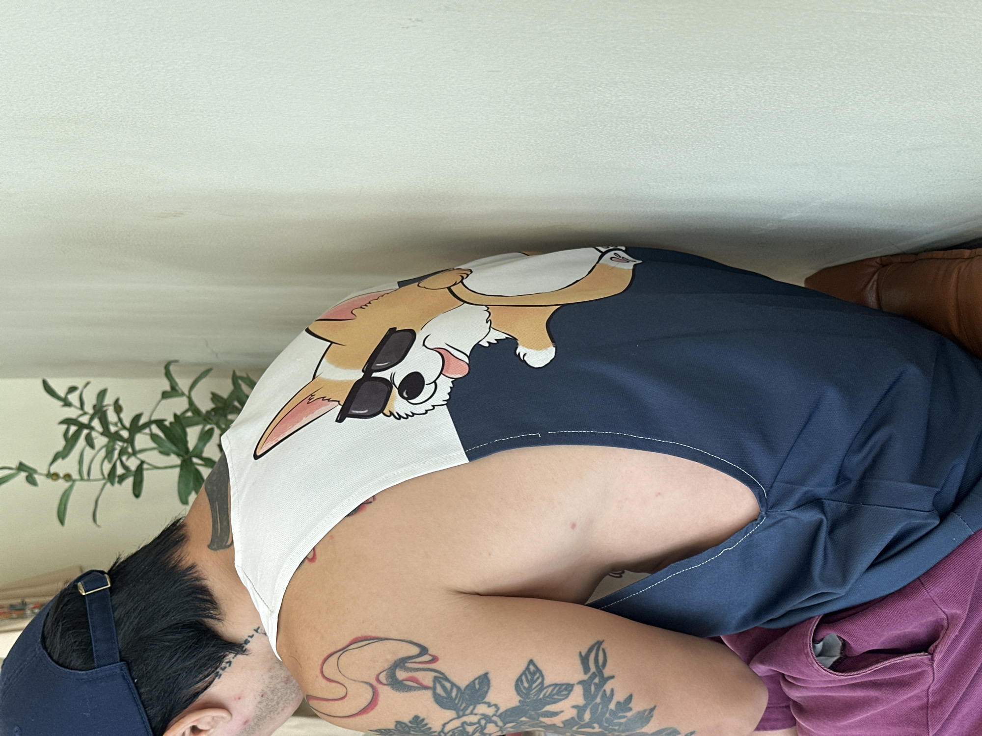 The image shows a person wearing a dark blue baseball cap, a white tank top with a corgi wearing sunglasses on the back, and dark blue shorts. The person has tattoos on their arm and neck. They appear to be sitting down. In the background, there's a plant and what appears to be a couch. The exact location in North Kuta, Indonesia cannot be determined from the provided information.
