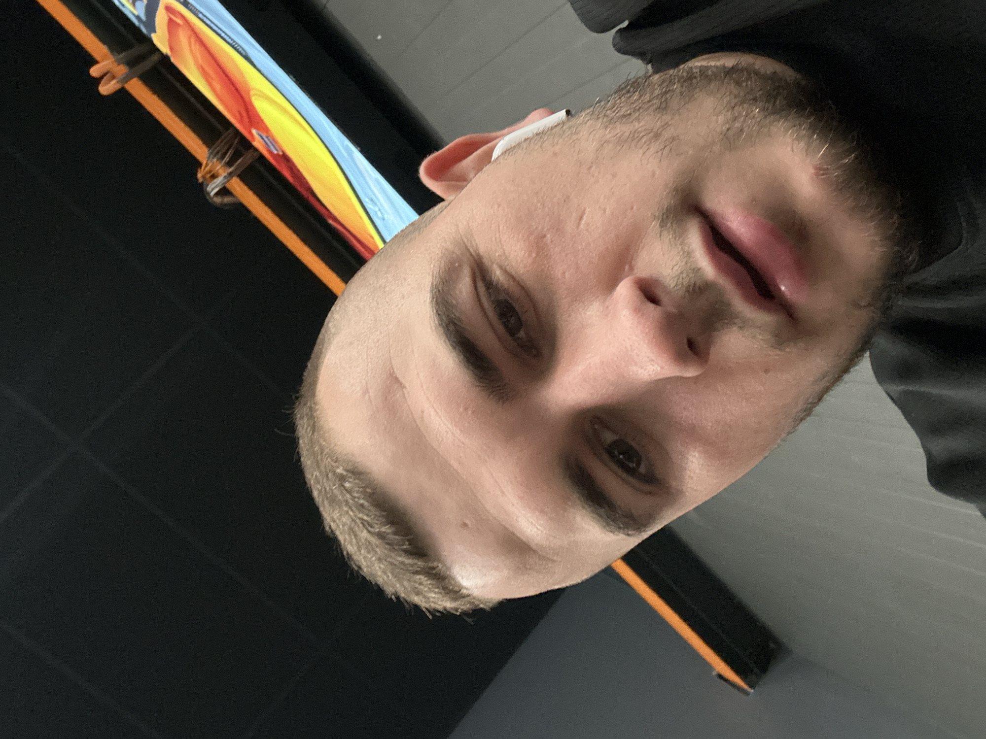The image is a close-up selfie of a man with short brown hair and a beard. He is looking at the camera with a serious expression. He appears to be wearing an earbud in his right ear. The background is out of focus but appears to be an interior space with a black ceiling and grey walls. There is a colorful banner with orange and yellow visible in the upper left corner. There is no evidence to determine the location of the subject in Plovdiv, Bulgaria.