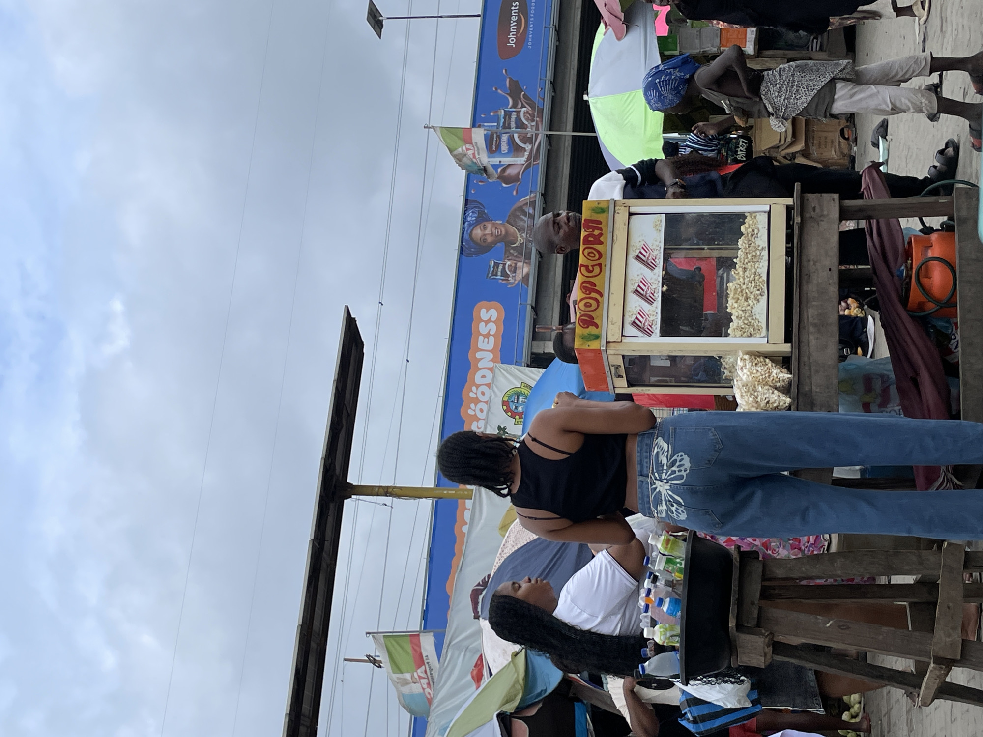An outdoor market scene unfolds under a cloudy sky in Lekki, Nigeria. A popcorn cart, its glass front displaying freshly popped kernels, is a central focus, operated by a man. To the left, a woman in a white t-shirt stands near a wooden stand laden with various bottles of beverages. Behind her, another woman, seen from the back, wears a black tank top and blue jeans with a butterfly design. A young child, adorned in a blue headscarf and draped clothing, stands to the right of the popcorn cart, appearing to interact with the vendor. Various stalls with umbrellas and a large blue billboard advertising "JOHNVENTS FOODS" and "GOODNESS" featuring a woman in traditional attire, are visible in the background. The ground is sandy or dusty, with scattered items suggesting a bustling marketplace environment.