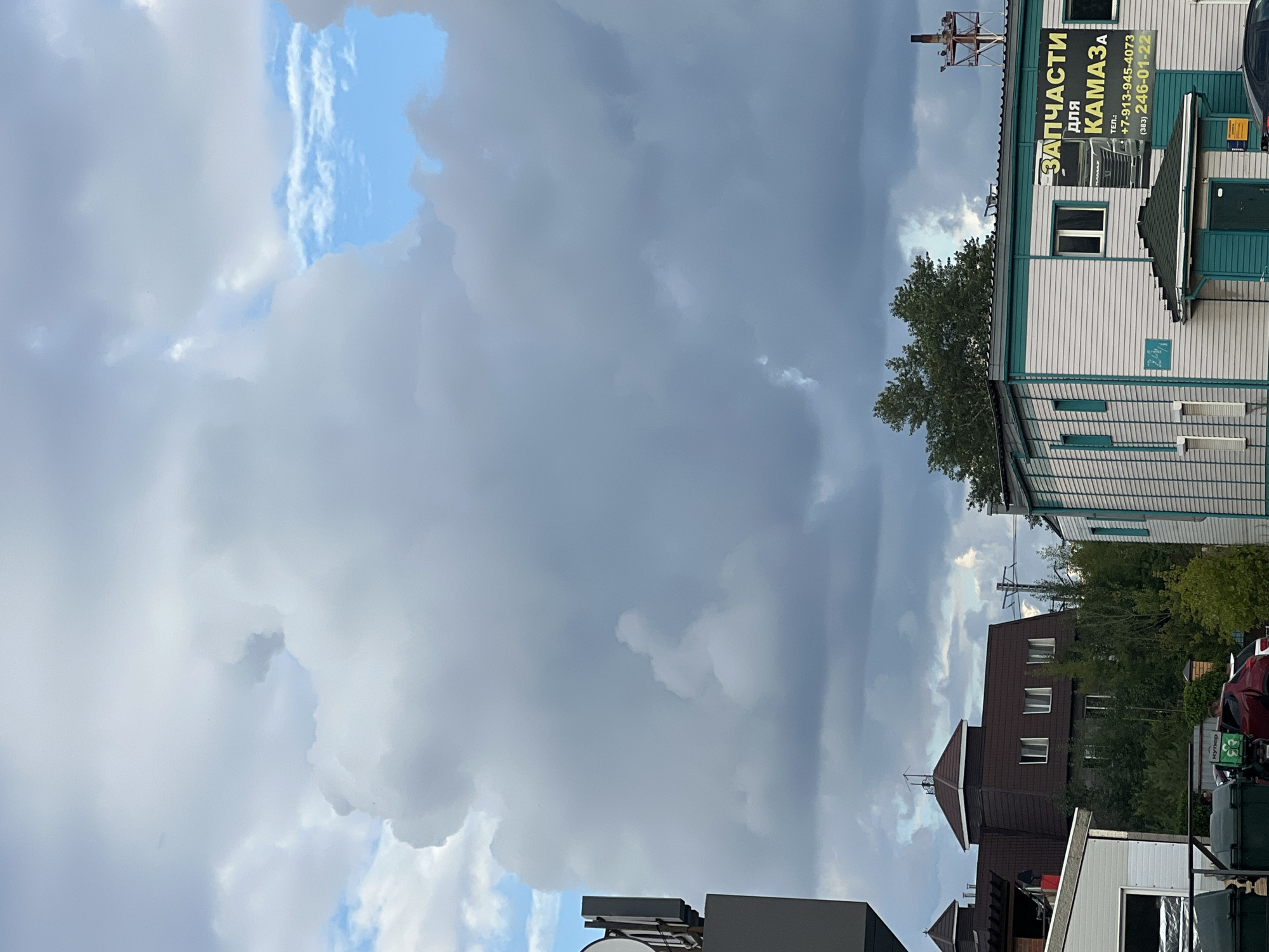 This image was captured from an upward-looking perspective, featuring a dramatic sky filled with heavy, gray clouds that partially obscure a bright blue sky. In the foreground, a modern building with white siding and teal trim stands under construction or renovation, with visible conduits and electrical wiring on its exterior. A sign on the building reads "ЗАПЧАСТИ ДЛЯ КАМАЗа" (Spare parts for KAMAZ) in yellow lettering, along with phone numbers. To the left, a dark structure, possibly a billboard or signpost, is partially visible. Further in the background, a multi-story building with a brown, steeply pitched roof and multiple windows is nestled among dense green trees. The overall setting appears to be an industrial or commercial area within the city of Novosibirsk, Russia.