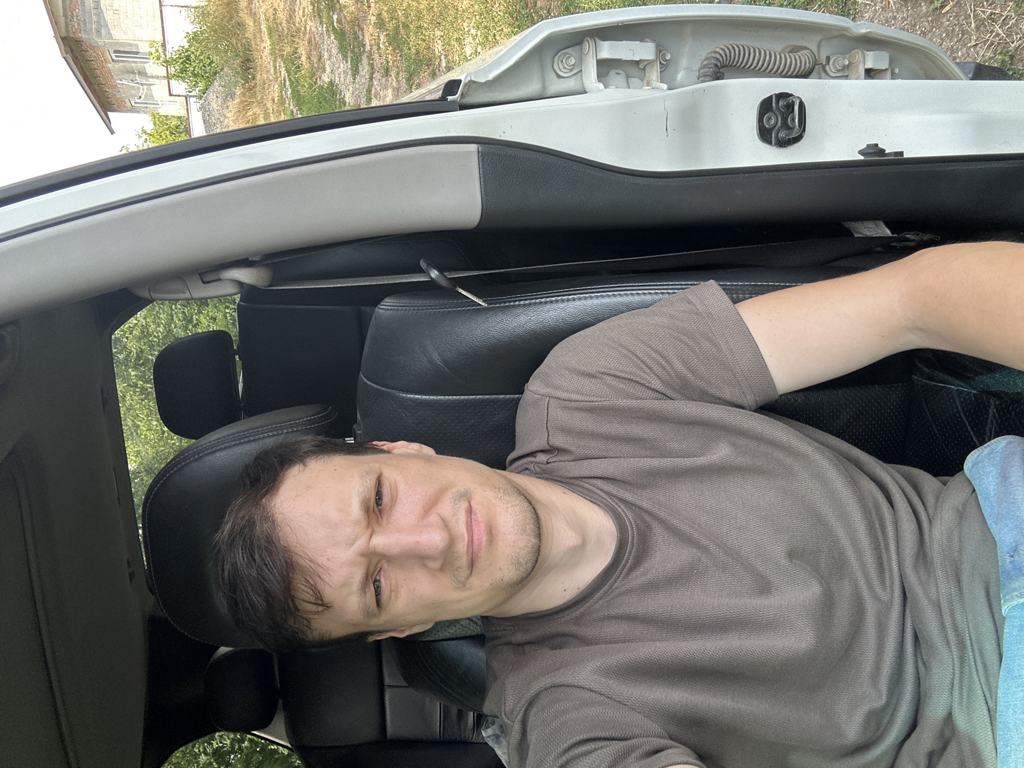 A man with short brown hair and a grimace is sitting in the driver's seat of a white car, looking at the camera. He is wearing a brown t-shirt and blue jeans. The car's interior is black leather. The driver's side door is open, revealing the exterior of the car and a rural setting with a brick building in the background. The image appears to be a selfie taken in Makiv, Ukraine.