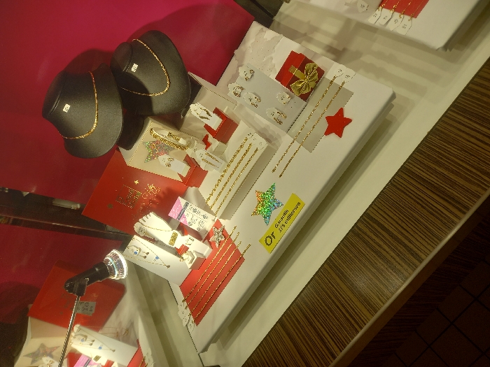 This image captures a jewelry display within a retail setting in Vendin-le-Vieil, France. The central feature is a white display platform showcasing various gold-colored jewelry items, including multiple necklaces, bracelets, earrings, and rings, presented on an assortment of white stands and forms. Two black, neck-shaped busts are positioned in the upper right quadrant, each exhibiting a gold-colored necklace and bearing a small white price tag. A directional spotlight with a silver-colored casing and a clear, faceted lens extends from the upper left, directly illuminating the display. The background consists of a solid magenta-pink wall. Decorative elements on the display include several red cardboard stars, a holographic star sticker, and a small red gift box adorned with a gold ribbon. A yellow label prominently displayed on the white platform reads "Or Garantie 375 millièmes" (Gold Guaranteed 375 thousandths). The immediate foreground consists of a dark brown counter featuring a distinct, striped wood grain pattern, and a dark tiled floor is partially visible in the bottom-left corner of the frame. No individuals are present in the scene.