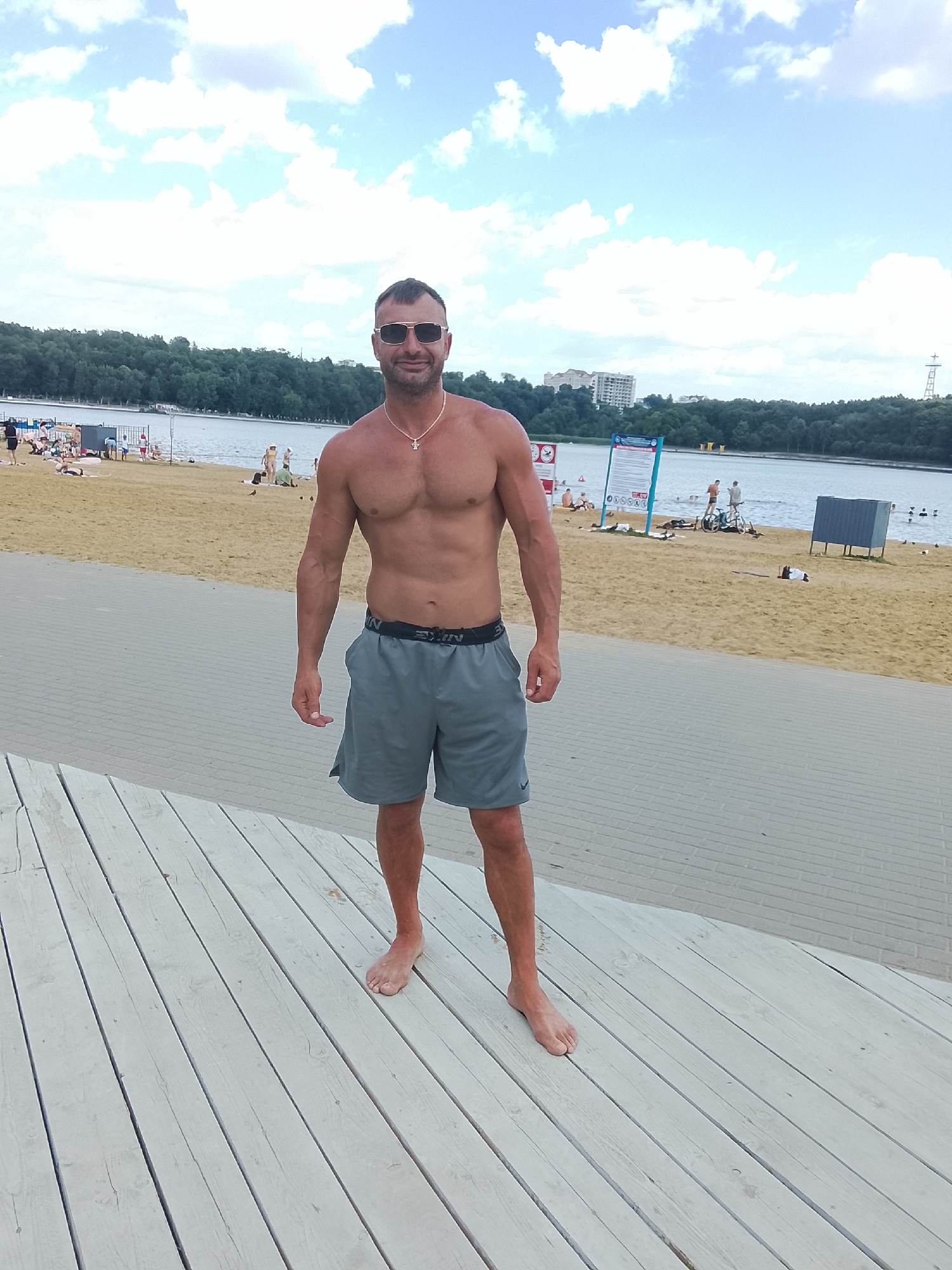 A man with a muscular physique stands on a wooden platform overlooking a beach and a body of water. He is wearing grey shorts and sunglasses, and a necklace with a pendant. The beach is populated with several people sunbathing, swimming, and standing. In the background, there are trees, buildings, and a clear sky with scattered clouds. The setting appears to be a recreational area, possibly a beach resort or a park with a lake. There is no information to suggest this location is in Chișinău, Moldova.