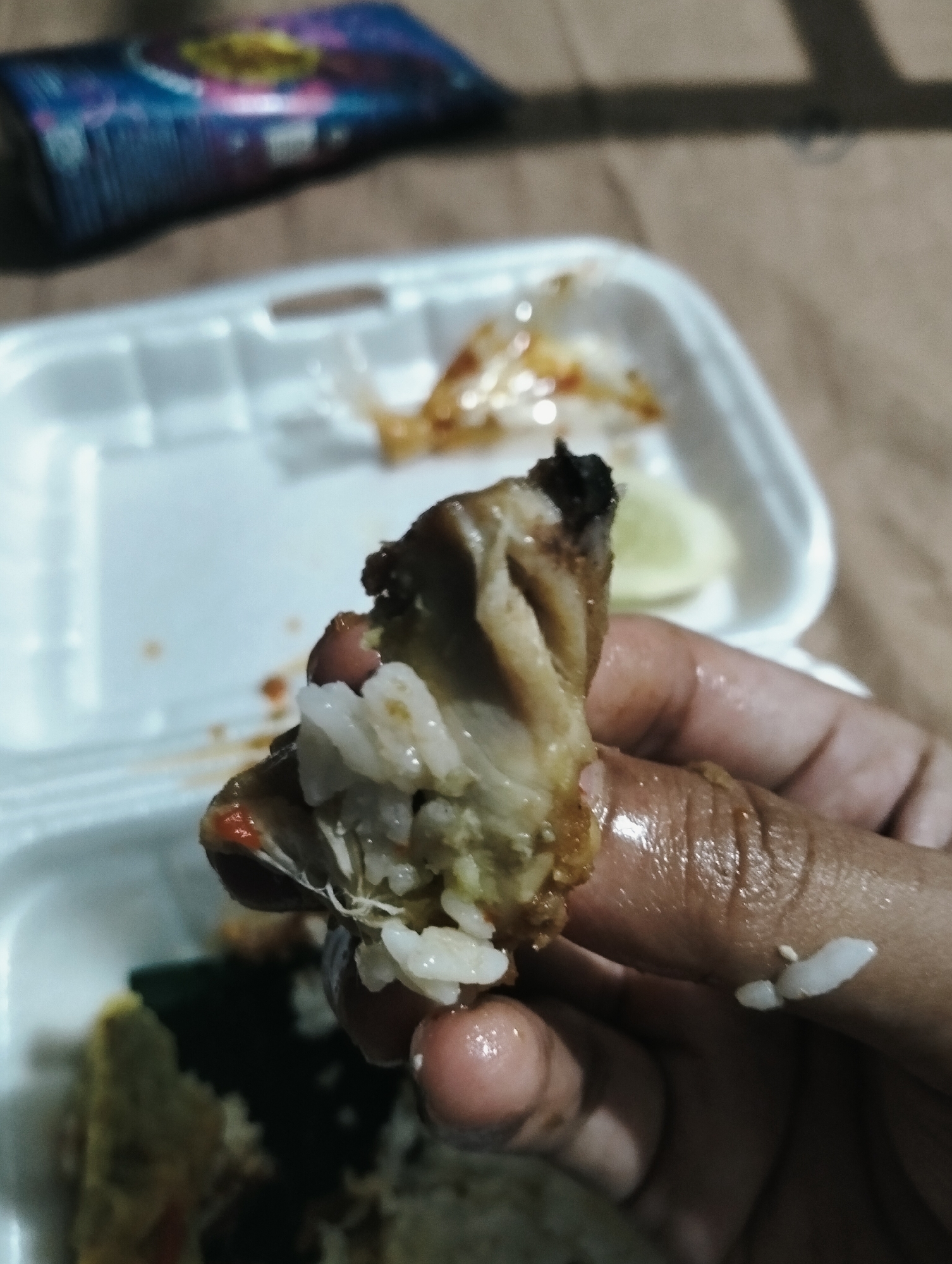 A close-up shot shows a person's hand holding a piece of fried chicken with rice. The chicken is dark brown and appears to be slightly burnt on one end, with some white rice stuffed inside. The hand is oily, and a few grains of rice are visible on the fingers. In the background, a white styrofoam food container is visible, with remnants of food and sauce. Partially visible in the upper left corner is a blurred image of a colorful package, possibly a snack or cigarette pack. The scene appears to be indoors, with a neutral-colored surface beneath the container.