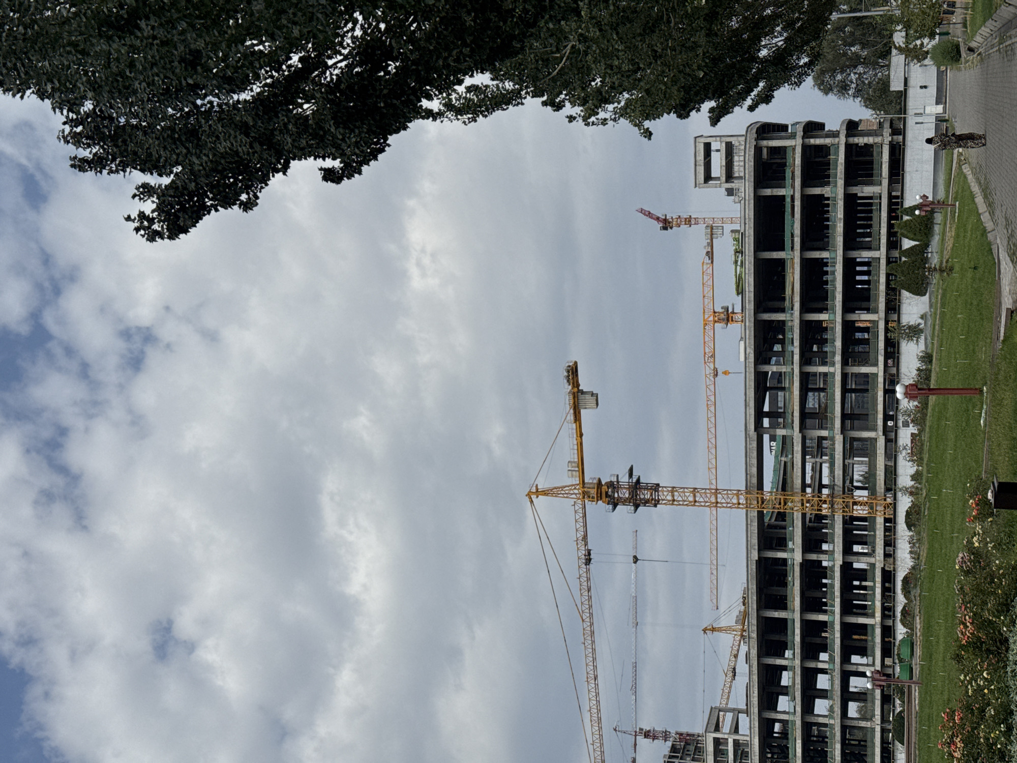 The image depicts a construction site in Bishkek, Kyrgyzstan, under a cloudy sky. A large, multi-story building is under construction, its concrete frame exposed. Several construction cranes, including a prominent yellow tower crane and a red one, are visible amidst the structure. In the foreground, a paved walkway winds through a grassy area with scattered trees and bushes. A person, possibly a woman, walks away from the camera on the walkway. A bicycle is parked nearby. The overall scene conveys an atmosphere of ongoing development within the city.