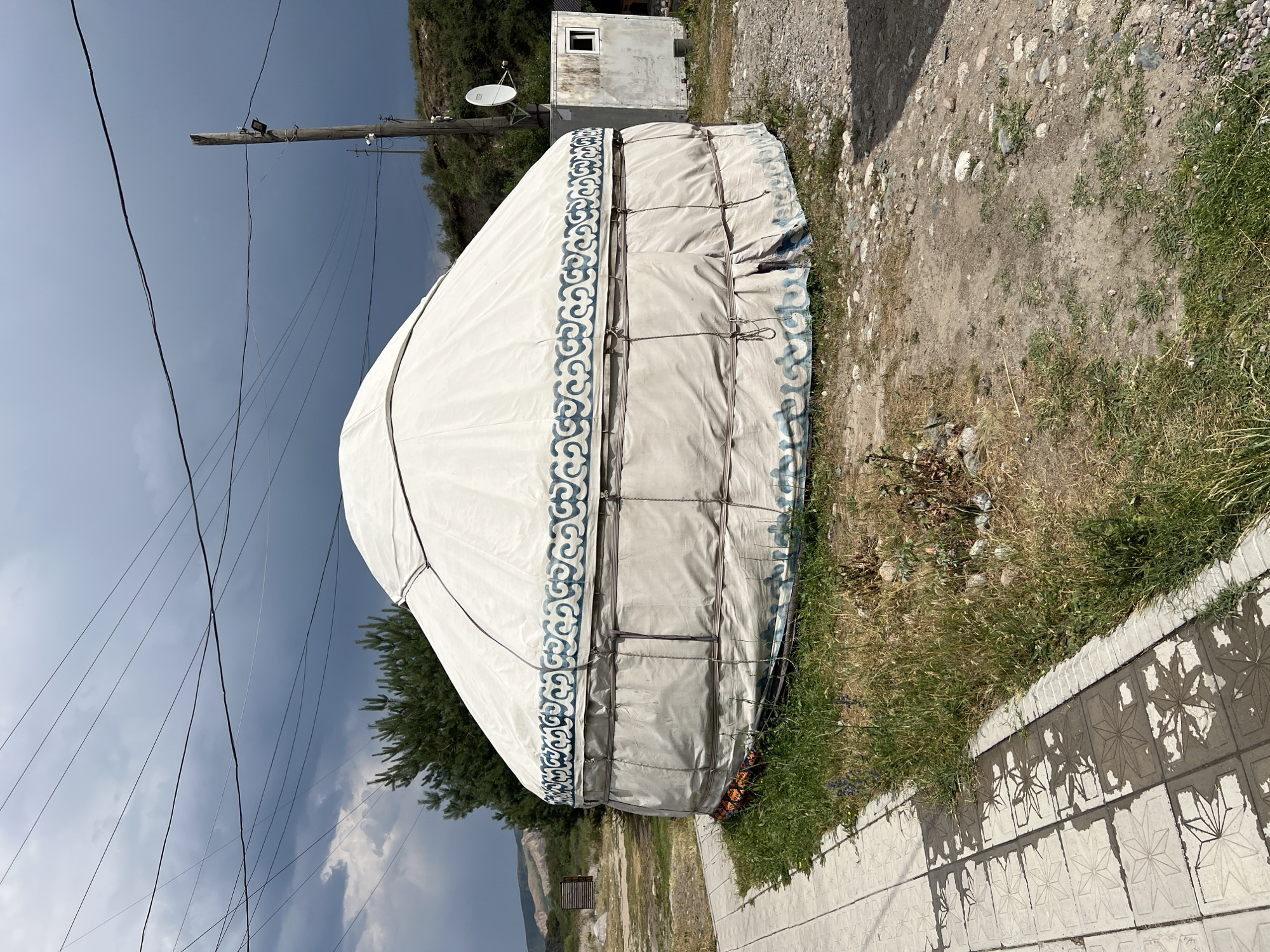 A white yurt with blue ornamental patterns sits on a patch of dry grass next to a tiled walkway. The yurt is circular with a domed roof and appears to be constructed of fabric stretched over a frame. A utility pole with electrical wires and lights stands to the right of the yurt, next to a small, weathered white building with a single window. In the background, a pine tree and a grassy hillside are visible under a dark, cloudy sky. The ground around the yurt is a mix of dirt, gravel, and sparse vegetation. The scene is likely located in Yuryevka, Kyrgyzstan, based on the presence of traditional yurts and the overall landscape. There are no people visible in the image.