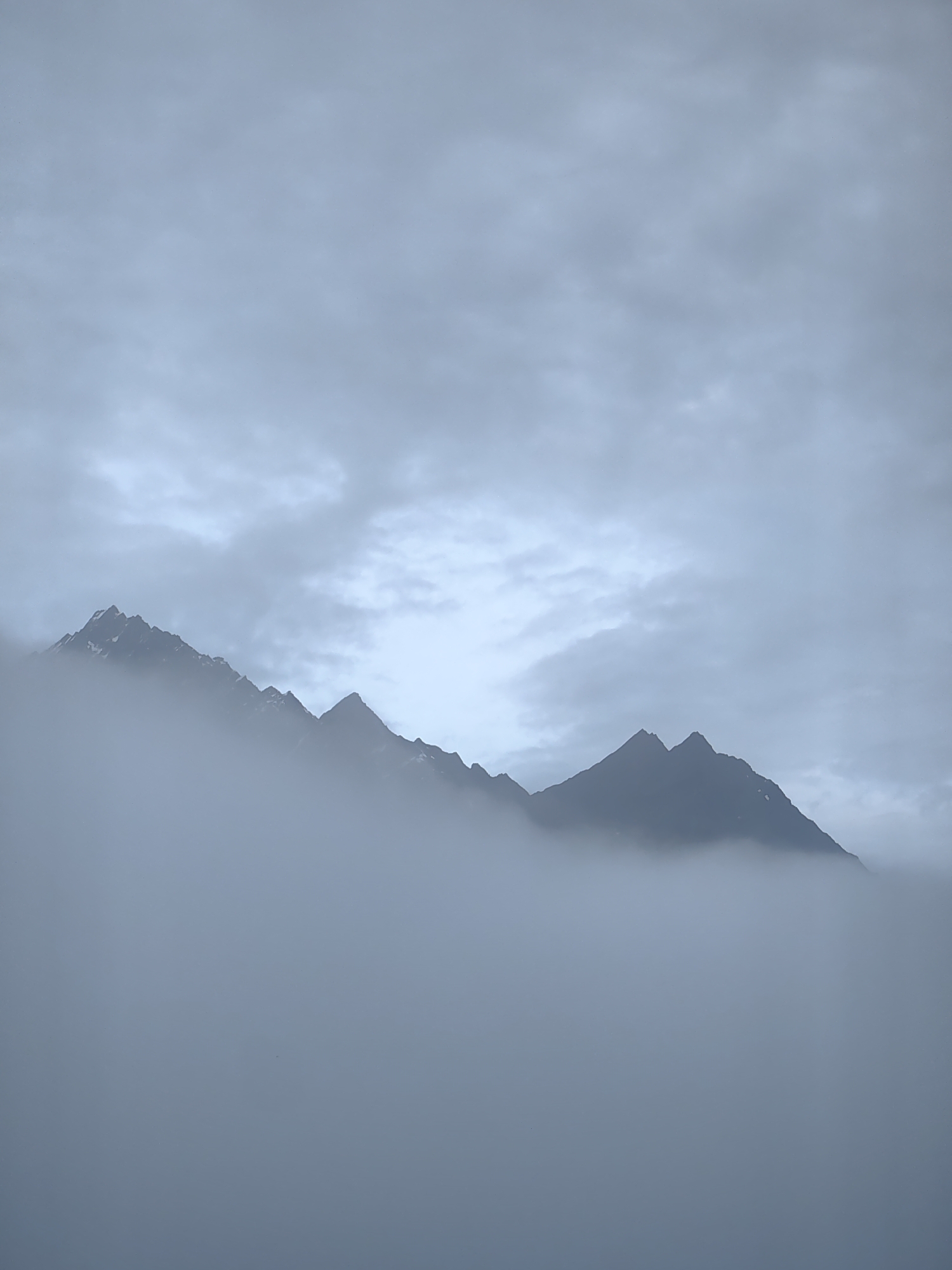 The image displays a mountain range shrouded in dense fog, with only the jagged peaks piercing through the mist. The sky above is overcast with heavy, gray clouds. The overall scene is monochromatic, dominated by shades of gray and blue, giving it a somber and ethereal atmosphere. The location is identified as Vashisht, India. There are no people, animals, or man-made structures visible in the image.