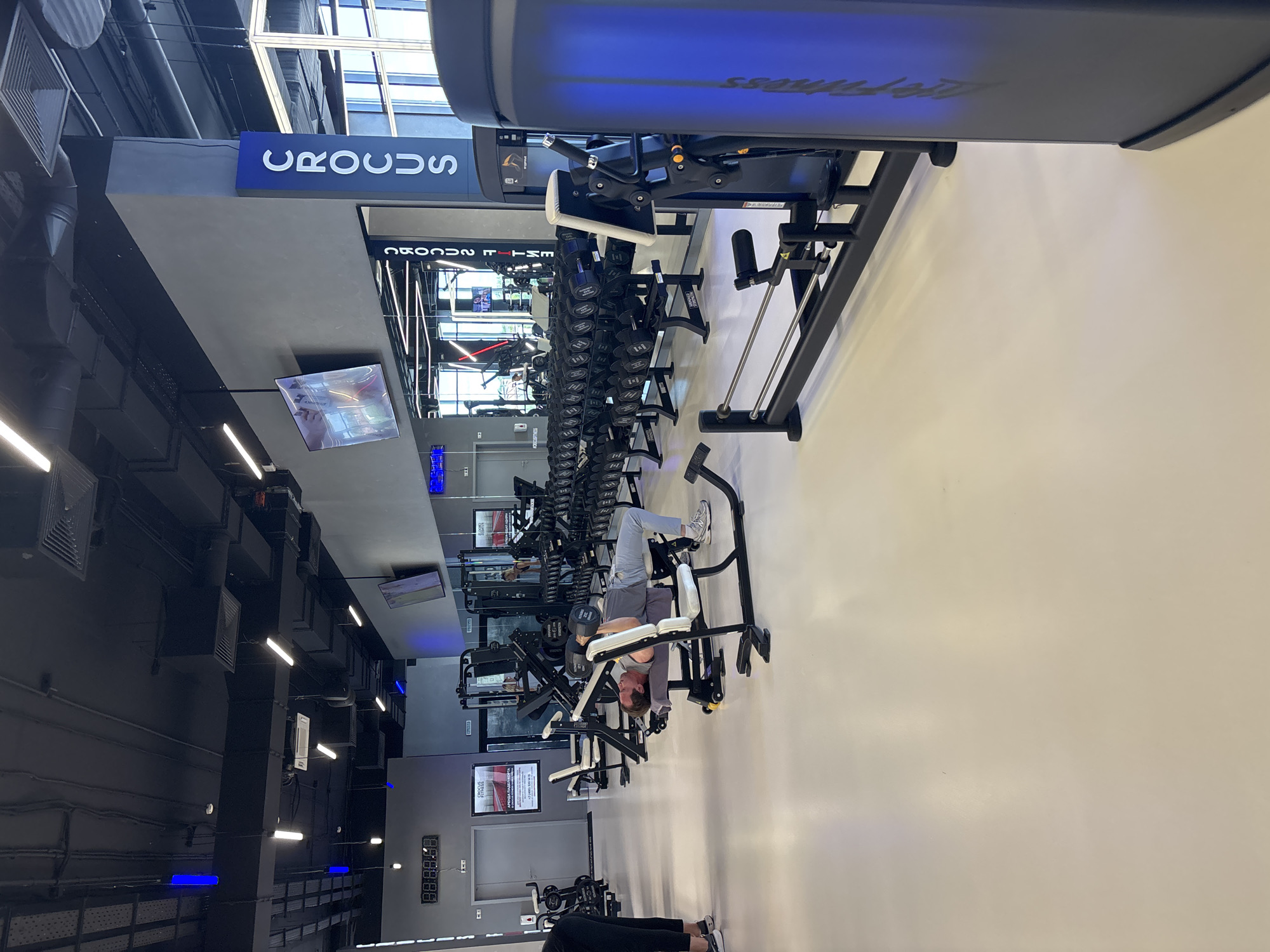The image shows the interior of a modern fitness center. In the foreground, a man is lying on a weight bench, performing a dumbbell press. He is wearing a grey t-shirt and light blue pants. To the left of the bench, a person in black athletic wear is partially visible, facing away from the camera. The gym features rows of dumbbells on racks, weight machines, and a clean, light-colored floor. A large mirror lines one wall, reflecting the gym equipment and ceiling lights. Overhead, the ceiling is dark and industrial, with visible pipes and ventilation systems, interspersed with bright fluorescent and blue LED lights. A prominent vertical sign with the word "CROCUS" in blue letters is visible on a pillar. The overall environment is well-lit and organized, typical of a commercial gym. The location is identified as Monolit, Russia.