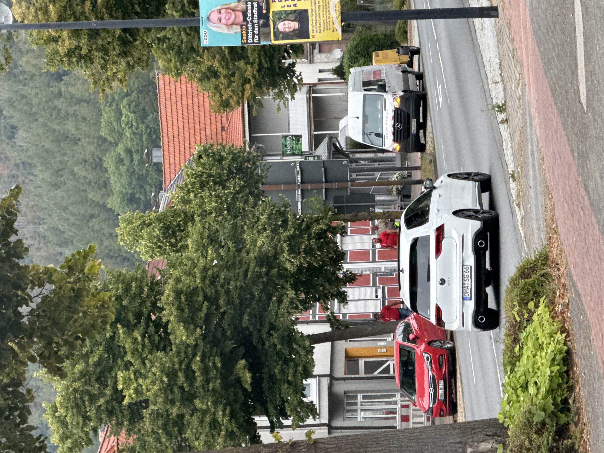 The image depicts a street scene in Bad Lauterberg im Harz, Germany, on a cloudy day. In the foreground, a white Volkswagen Golf GTI is driving on the right side of the road, with its rear facing the camera. To its left, a red Opel Astra is parked. Two men in red shirts are visible standing near the parked car, and another man in a light brown shirt is standing further down the sidewalk. A white Mercedes-Benz Sprinter van is driving on the opposite side of the road, towing a yellow trailer. On the right side of the image, a lamppost supports two posters: the top poster features a woman identified as "Saskia Dittrich-Cziolles für den Stadtrat", and the bottom poster has an image of a woman and some German text. The background shows buildings with red-tiled roofs and a dense forest covering a hillside. Lush green foliage lines the sidewalk in the lower left of the frame.