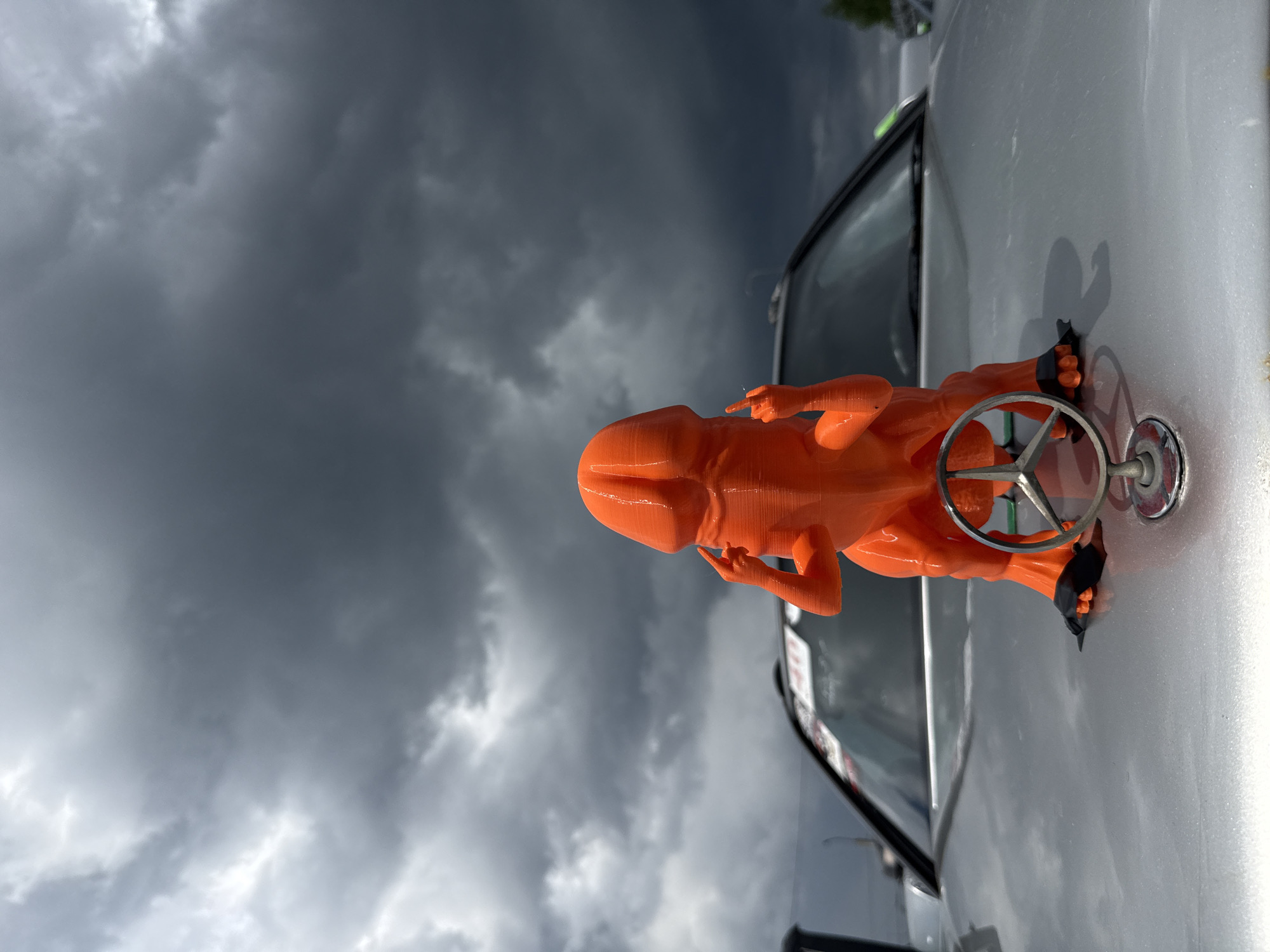 A low-angle shot shows a 3D-printed orange figure positioned on the hood of a silver vehicle. The figure appears to be seated and grasping a circular emblem with a Mercedes-Benz logo. The car, partially visible, includes a windshield and a small portion of a side window. The backdrop showcases a dramatically overcast sky, filled with dark, foreboding storm clouds. The location is Unknown, Unknown.