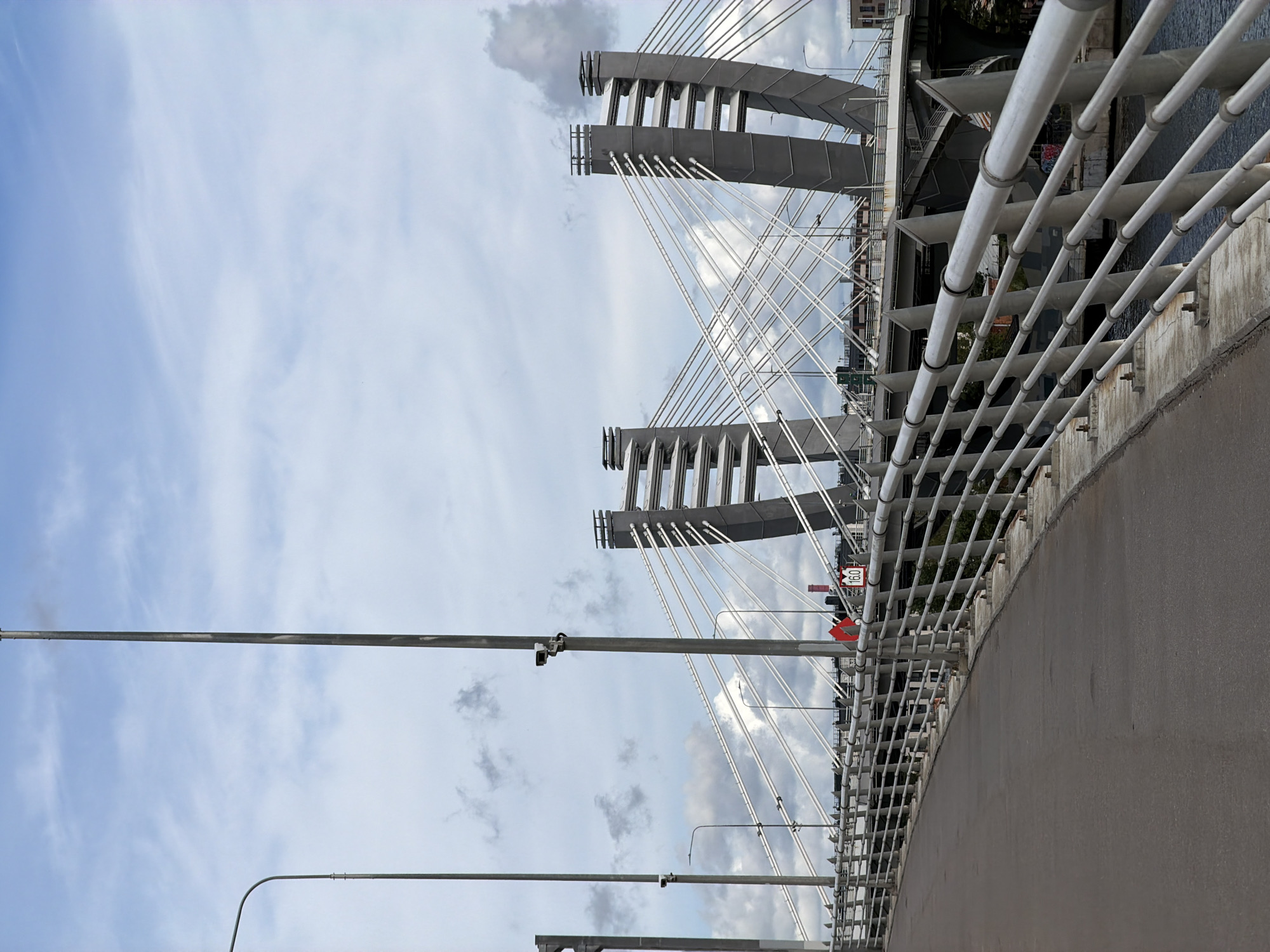 This is a photograph of a cable-stayed bridge in Saint Petersburg, Russia, under a cloudy sky. The perspective is from the pedestrian walkway on the bridge, looking forward. The walkway is paved with asphalt and bordered by a silver metal railing. In the background, two large, grey, angled pylons rise, from which numerous white cables extend to support the bridge deck. Additional streetlights and security cameras are visible on poles along the walkway. In the distance, other structures and buildings can be seen. A traffic sign displaying "16.0" is mounted on the railing. The water of a river or canal is visible below the bridge.