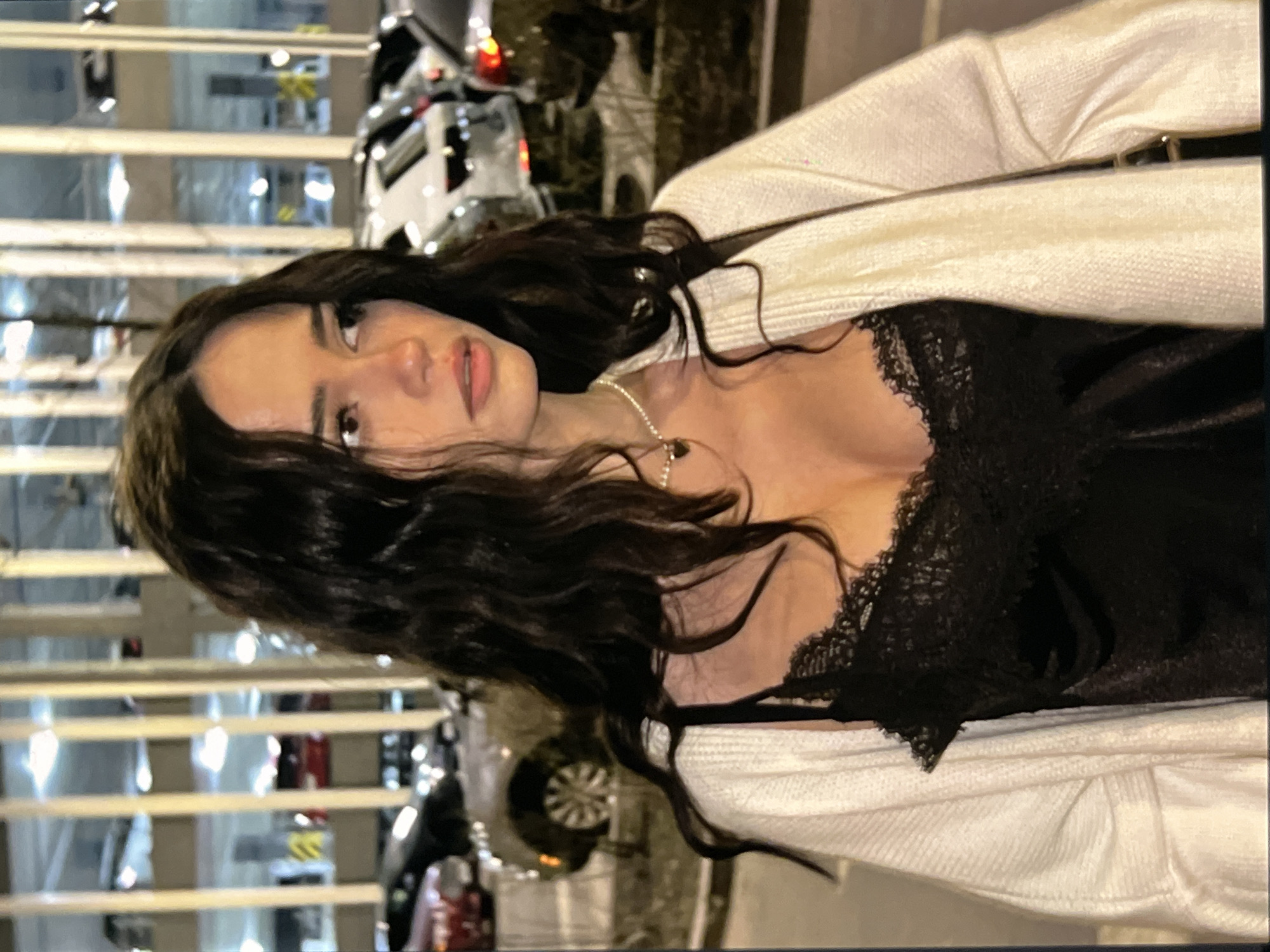 A young woman with long, dark, wavy hair stands outside a parking garage at night. She is wearing a black lace-trimmed camisole and a white knit cardigan. A silver necklace with a heart-shaped pendant is visible around her neck. In the background, cars are parked in the multi-level garage, illuminated by overhead lights. The woman is looking to her right with a thoughtful expression.