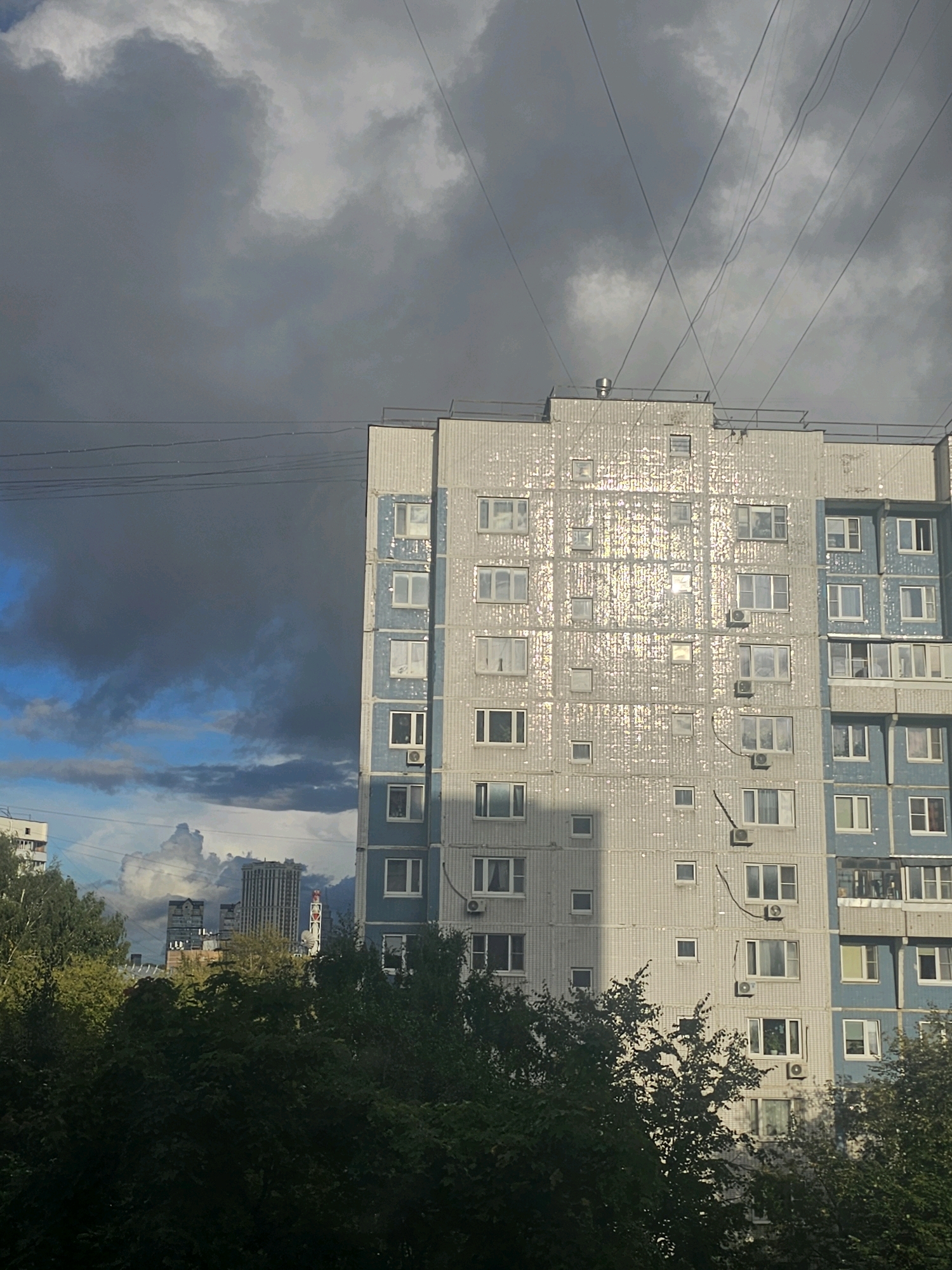 The image depicts a view of residential buildings and trees against a dramatic sky. The primary focus is a light-colored, multi-story apartment building with blue accents, its facade covered in reflective tiles that catch the sunlight. Numerous windows, some with air conditioning units, are visible on the building. To the left, another, lighter colored apartment building is partially visible, also with windows. In the distance, several taller modern buildings, including what appears to be a skyscraper, can be seen. The sky is a striking contrast, with dark, heavy clouds dominating the upper left portion of the frame, juxtaposed with areas of bright blue sky and lighter, more textured clouds on the right and lower portions. Thick green foliage from trees forms a dense border across the bottom of the image, partially obscuring the lower levels of the buildings. Several wires are strung across the upper portion of the frame, crossing over the buildings and sky. The overall scene suggests an urban environment, likely in Khimki, Russia, given the architectural style of the apartment buildings. There are no people or animals visible in the image.
