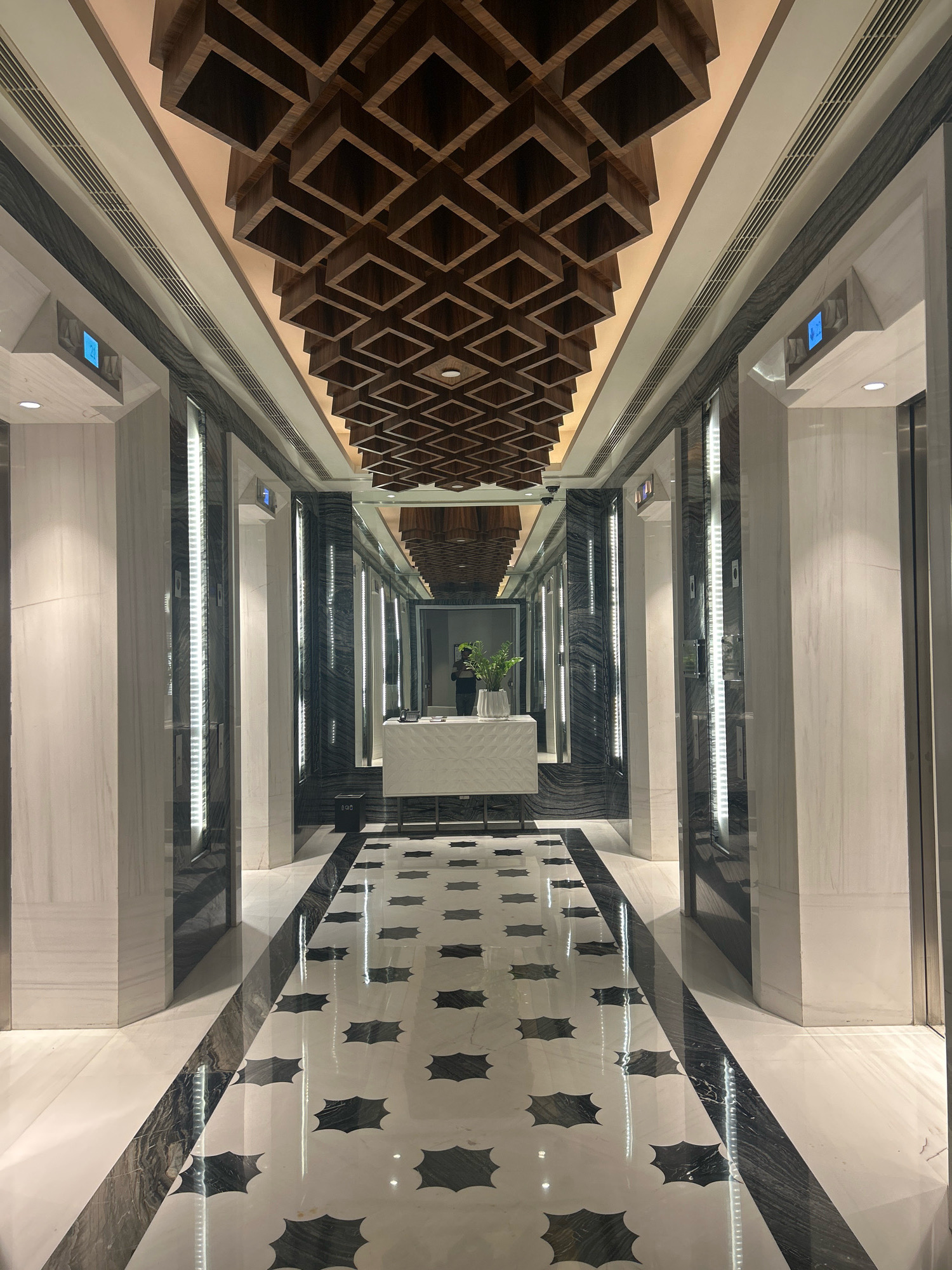 The image shows a long, well-lit corridor with polished marble floors featuring a black starburst pattern. On either side of the corridor are elevator doors, each with vertical LED lighting. The walls are a mix of white marble and dark, patterned stone. The ceiling is a distinctive design, with a grid of dark wooden squares recessed into a lighter surface, and subtle indirect lighting. In the distance, at the end of the corridor, there is a reception desk covered with a white cloth, with a potted plant on it. A person can be seen in the reflection of the far wall. The overall impression is of a modern and luxurious interior. Based on the visual cues, this location is likely a high-end hotel or commercial building in Hyderabad, India, given the prevalent architectural styles and materials often found in such establishments in the city.