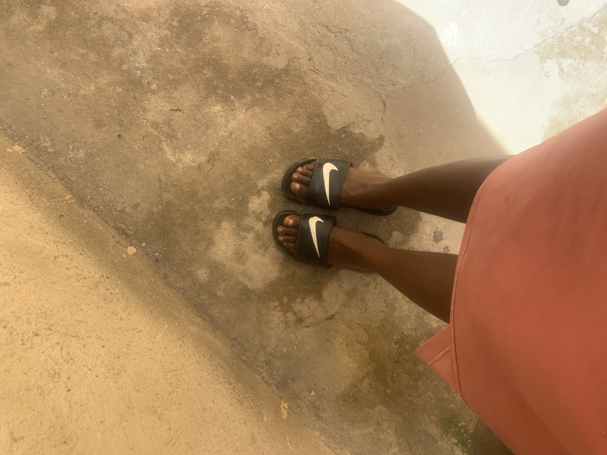 A top-down, medium-angle shot shows a pair of feet wearing black Nike-branded slides on what appears to be a weathered concrete surface. The legs are dark-skinned, and a portion of an orange garment is visible in the lower right of the frame. The background suggests an outdoor setting, possibly a street or courtyard. The scene appears to be in Rafin Par, Nigeria.
