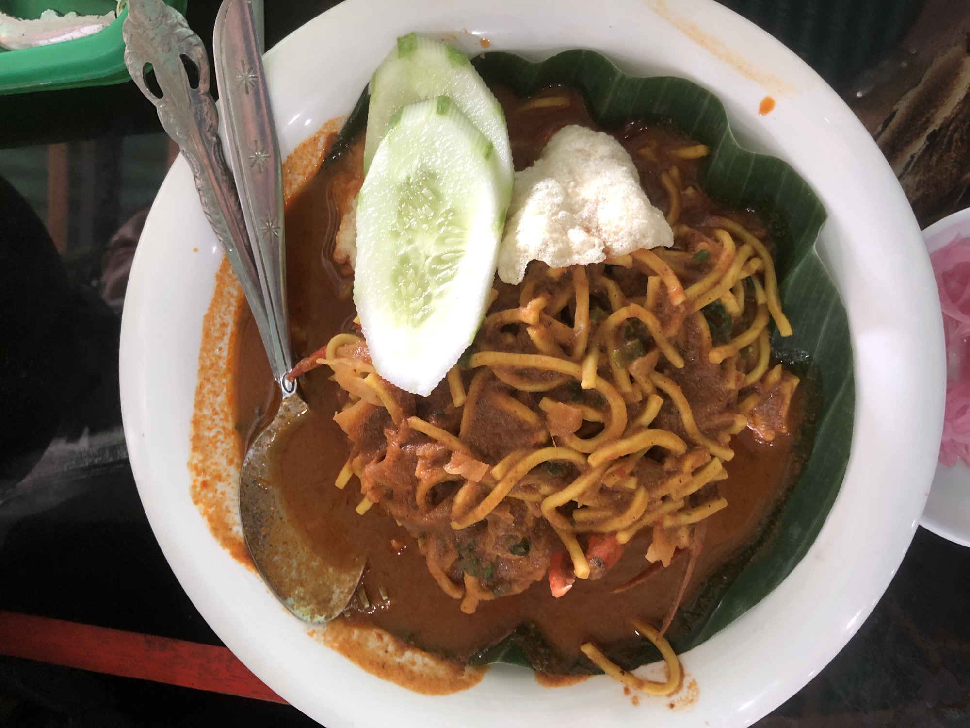 A top-down, eye-level, medium shot captures a plate of Mie Aceh, an Acehnese noodle dish, in Lancok, Indonesia. The dish consists of thick yellow noodles in a brown, curry-like broth, garnished with slices of cucumber, a cracker, and cilantro. It's served on a white plate lined with a banana leaf. Silverware rests on the edge of the plate. A small dish containing pickled onions can be seen near the edge of the image. The image is taken indoors, the location is a restaurant in Lancok, Indonesia.