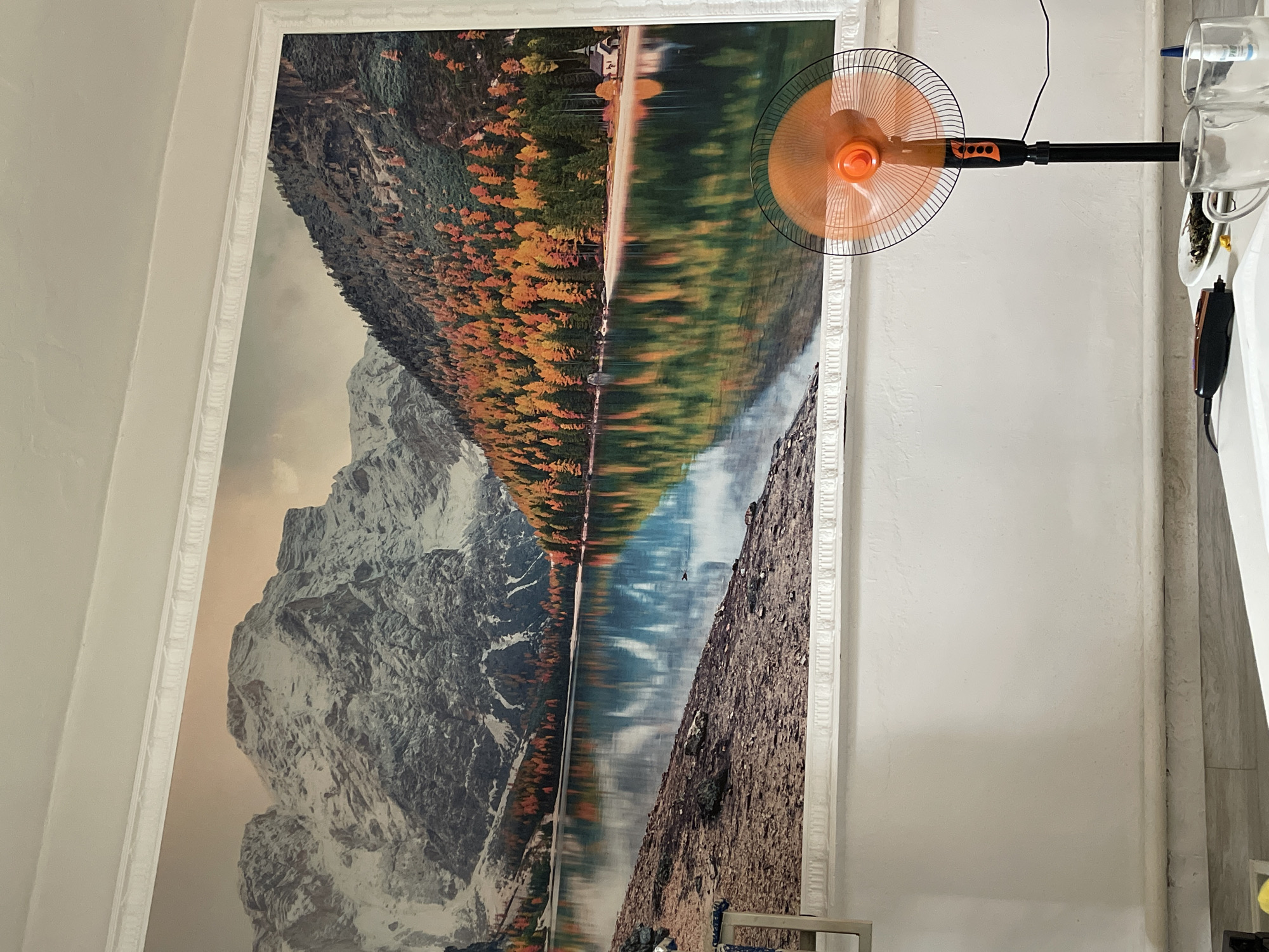 A large mural depicting a mountain lake scene with autumn foliage is mounted on a white wall. The mural features snow-capped mountains, a calm body of water reflecting the trees and mountains, and a small building on the far shore. In the foreground, an orange pedestal fan stands on the floor to the right of the mural. A white surface, possibly a table, is visible in the lower portion of the image, with some objects on it, including two clear mugs, a plate with food, and an electric grooming device. The flooring appears to be a light-colored material with a subtle pattern. The environment appears to be an indoor setting. The image does not provide any information about the city of Bishkek, Kyrgyzstan.