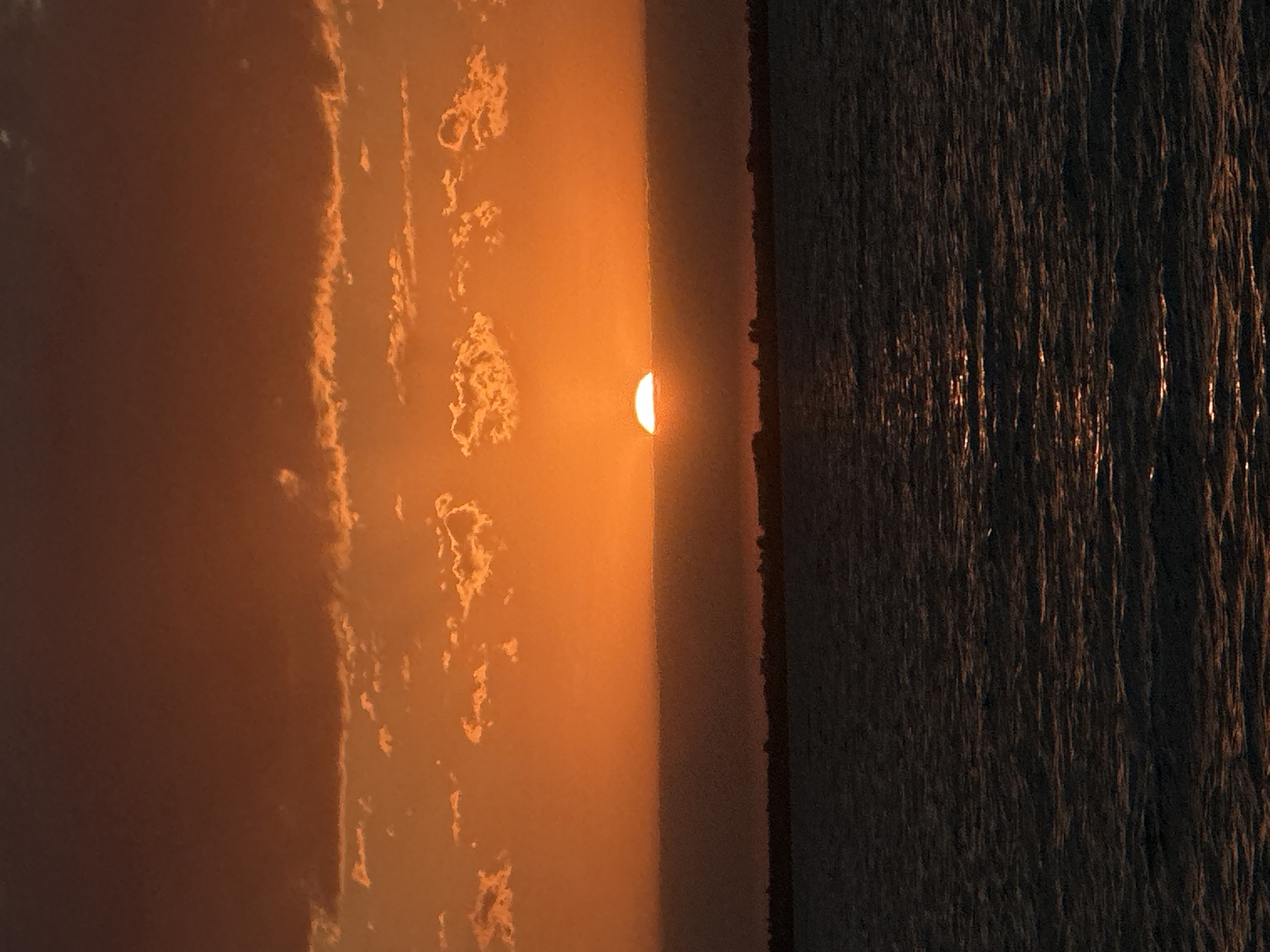The image depicts a sunset over a body of water, likely a lake or sea, with a distant shoreline visible. The sun is partially obscured by a band of clouds on the horizon, casting a warm orange glow across the sky and reflecting on the water's surface. The sky transitions from a deep orange at the horizon to a darker, reddish-brown hue at the top of the frame. Scattered clouds, illuminated by the setting sun, are visible in the middle section of the sky. A thin, dark band of clouds or haze sits directly above the horizon, behind which the sun is setting. The water is dark and appears somewhat choppy, with streaks of light from the sun's reflection. The distant shoreline is silhouetted against the brighter sky, showing the faint outlines of trees or vegetation. The image is taken from a low vantage point, looking across the water towards the sunset. There are no people or man-made structures clearly visible in the immediate foreground. Given the context, this scene could be observed from a waterfront area in Kokshetau, Kazakhstan, potentially along the shores of Lake Kokshetau, a prominent natural feature of the city.
