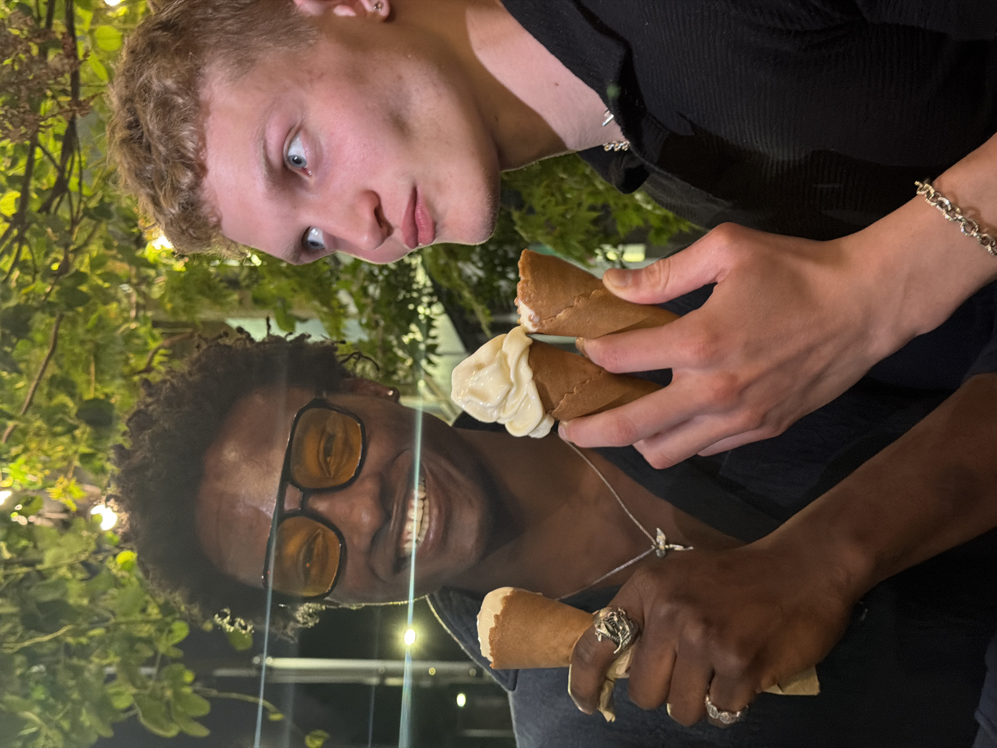 Two individuals are holding ice cream cones. One person has fair skin, short blonde hair, and blue eyes. The other person has dark skin, curly hair, and is wearing sunglasses. Both are wearing dark-colored clothing. The background includes greenery. The location appears to be an outdoor setting in the city of London, United Kingdom.
