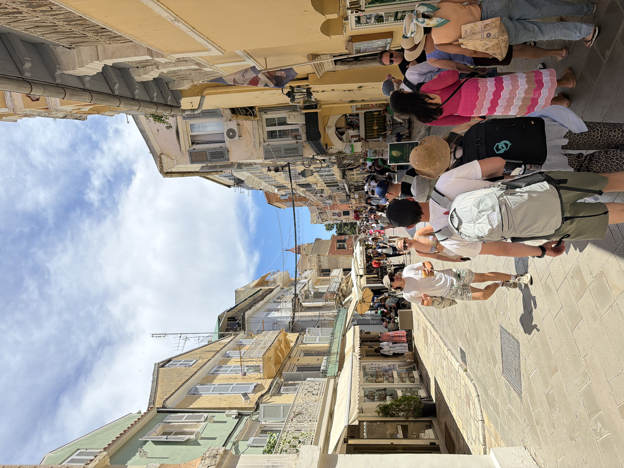 This image captures a vibrant street scene in Corfu, Greece, on a bright, sunny day. The perspective looks down a narrow, bustling street lined with multi-story buildings displaying traditional architecture. The sky, a clear blue with scattered white clouds, is visible above the rooftops of the colorful structures.
Numerous people, likely tourists, fill the street, walking and exploring. They are dressed in casual summer attire, including shorts, t-shirts, dresses, and hats, suggesting warm weather. In the foreground, a man in a white t-shirt and patterned shorts, wearing sunglasses, walks while holding a drink. Nearby, a woman in a white outfit and sunglasses is also visible. Further back in the crowd, individuals carry backpacks, one light grey and another black with a teal logo. A woman in a distinct pink and white striped skirt or dress stands amidst the other pedestrians. Some people are seen sitting at outdoor tables belonging to establishments further down the street.
The buildings flanking the street are painted in a mix of pastel colors, including yellow, light green, and light pink, with some appearing weathered. They feature numerous windows, many with closed shutters, and ornate balconies with decorative railings. Air conditioning units and exterior pipes are visible on some walls. A Greek flag hangs prominently from one of the buildings. Shopfronts with display windows and awnings line the street, indicating retail activity. A notable green sign with an olive branch logo clearly reads "THE CORFU OLIVE OIL SOAP FACTORY," confirming the location context. The street is paved with light-colored flagstones, and drain covers are visible. The bright sunlight and the casual clothing indicate it is daytime, likely during a warm season, contributing to the lively and active atmosphere of this popular tourist destination.