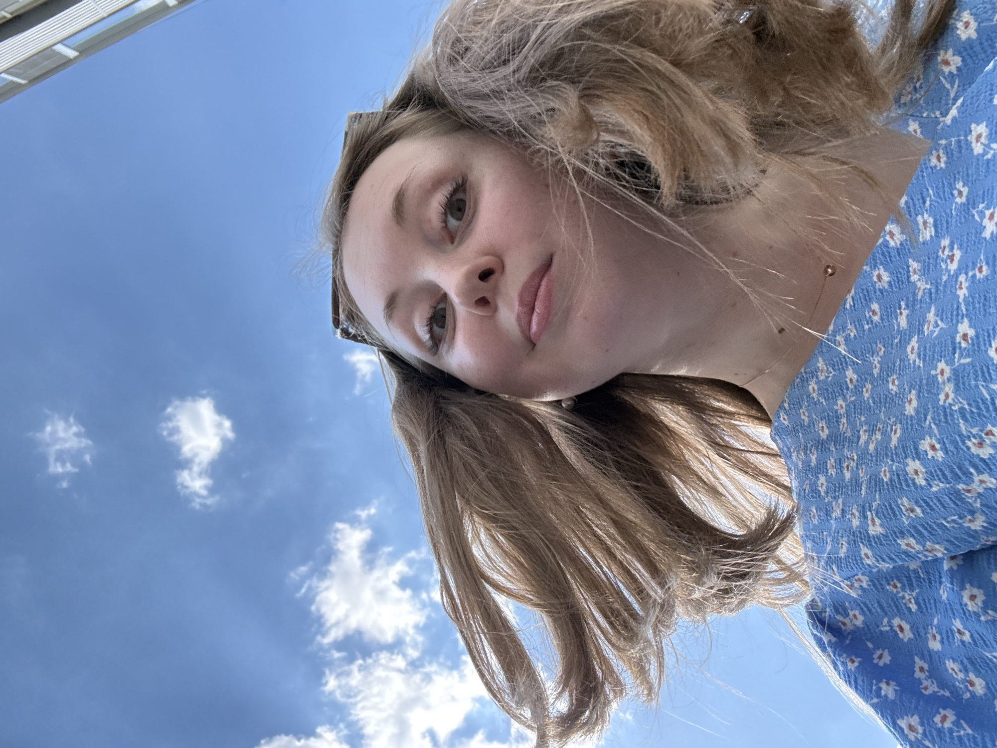 A low-angle, off-center selfie shows a young woman with light brown hair and fair skin, wearing a blue floral print dress, against a partly cloudy blue sky. The woman's face is tilted towards the camera, her expression neutral. Part of a structure, possibly a building, is visible in the upper left corner. The location is Unknown, Unknown.