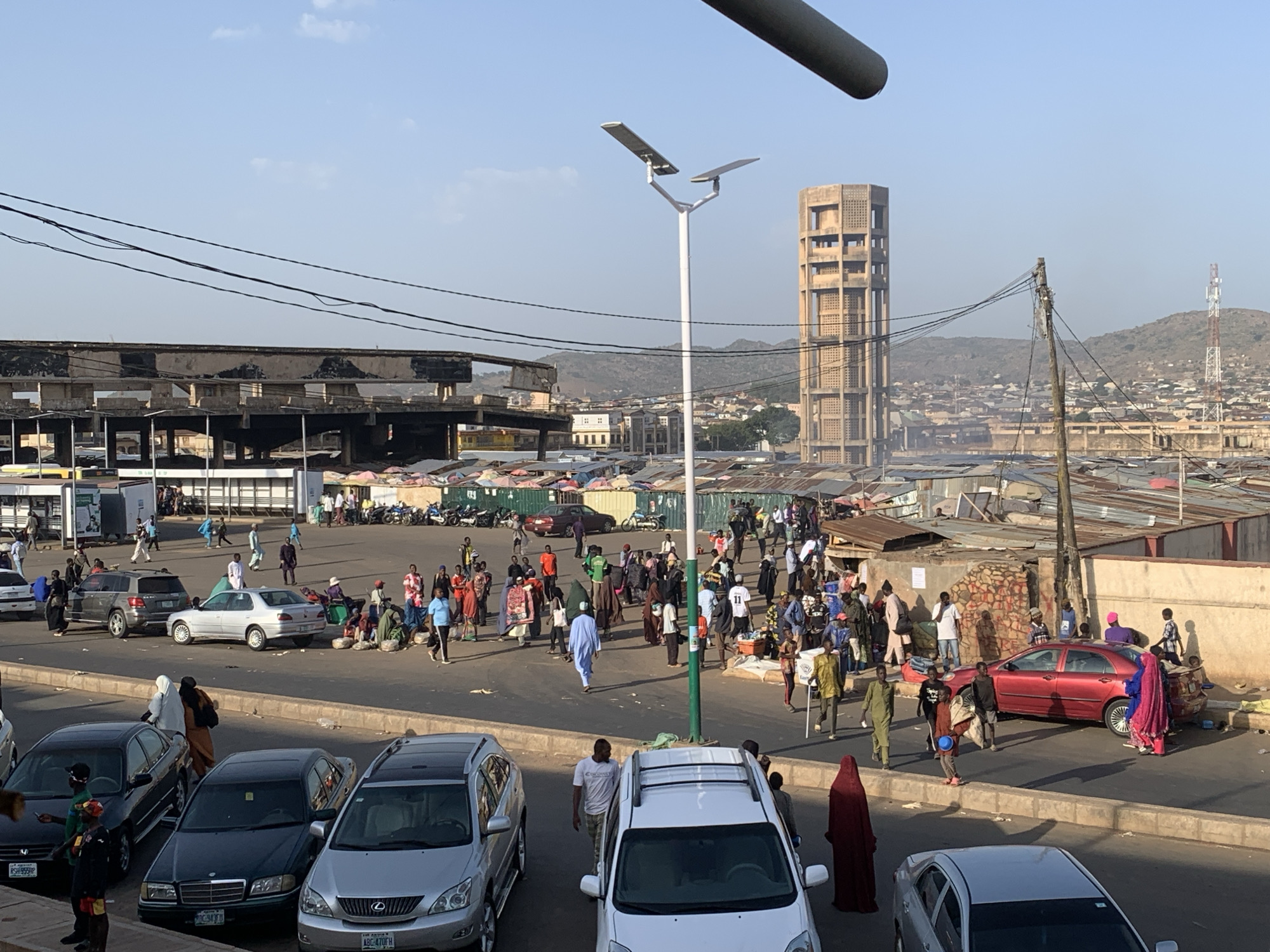 The image depicts a bustling market scene in Jos, Nigeria. In the foreground are several parked cars. A busy roundabout features people walking, some carrying items or wearing traditional clothing. In the background, there's a concrete tower with square cut-outs and a hillside with dense housing. Overhead power lines run across the scene. A large partially constructed building sits in the middleground to the left, while the right foreground has a street market with stalls and vendors. A solar powered lamp post is next to a group of people. The sky is blue with some light haze.