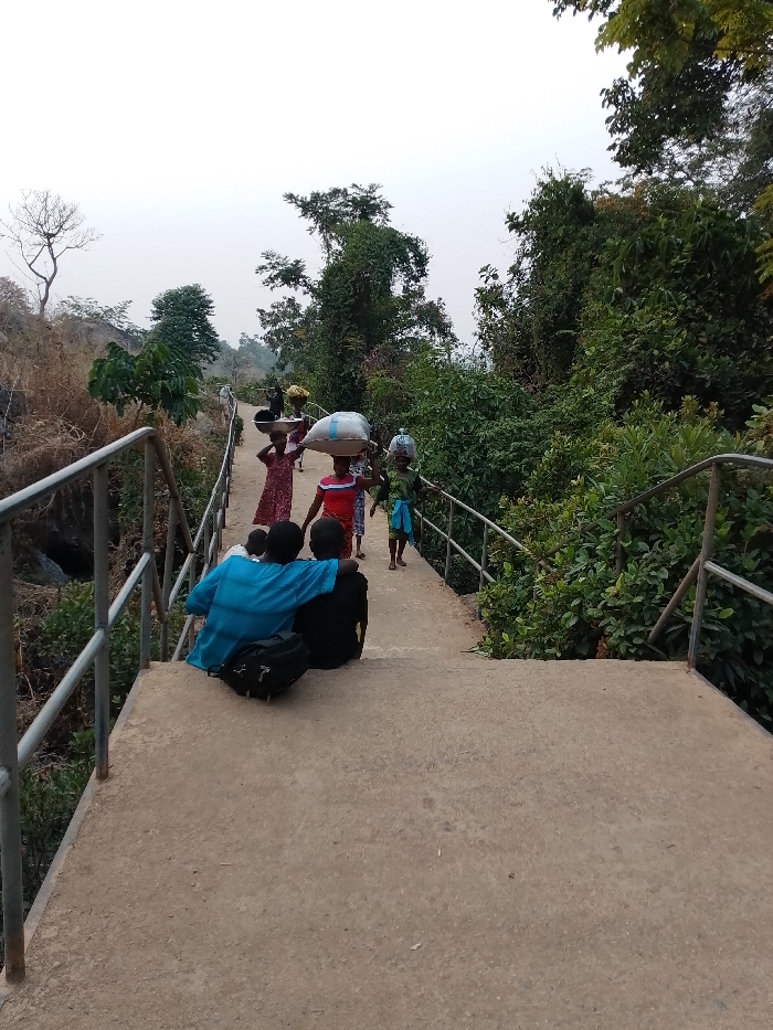 A bridge spans a lush, green ravine. Women carry items on their heads, walking across the bridge. Two individuals sit with their backs to the viewer. One wears a blue shirt and leans toward another individual with a black shirt, a backpack resting beside them. The bridge has metal railing on both sides and leads into a dense forest area. The sky is overcast, creating a soft, diffused light.