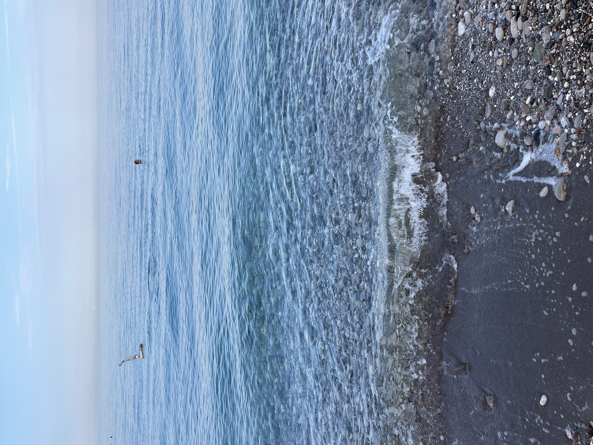 A person is swimming in the calm, blue waters of the Adriatic Sea near the city of Bar, Montenegro. In the distance, a floating object with a striped pattern is visible on the water. The shore is covered in smooth pebbles and dark sand, with gentle waves lapping at the edge. The sky is a pale blue with a few wispy clouds, and the horizon is hazy.