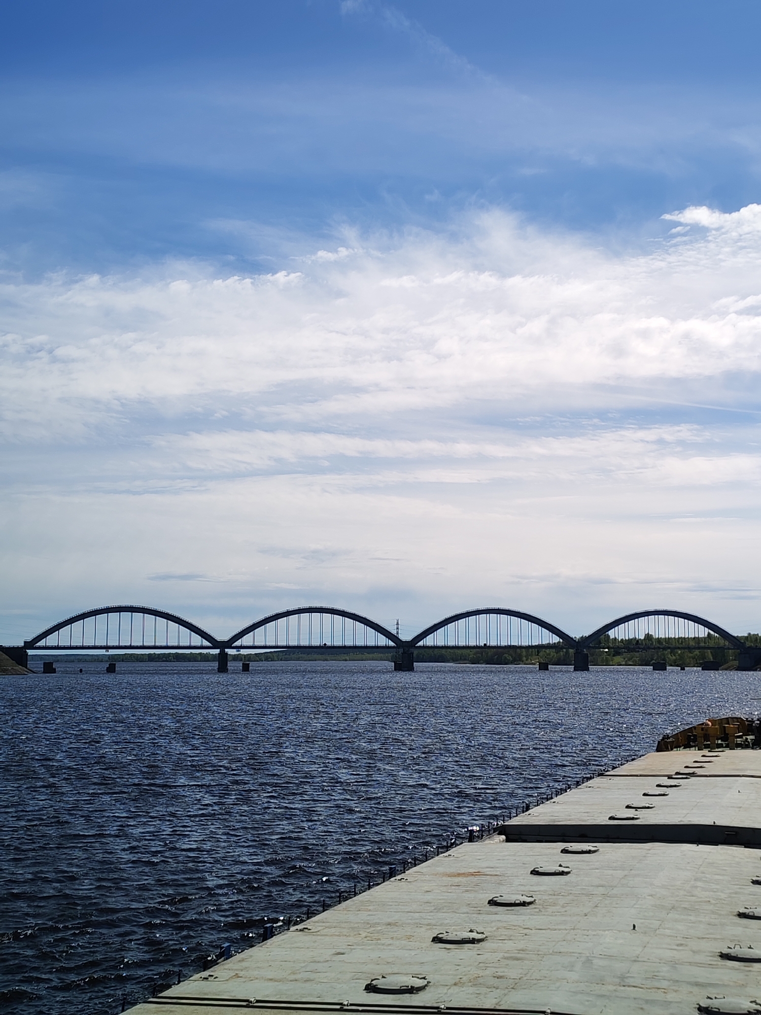 The image captures a wide-angle view of a bridge spanning a body of water in Krasnyi Rybak, Russia. The bridge features multiple arched spans, supported by pillars submerged in the water. The water's surface is rippled, indicating a slight breeze. The sky above is a mix of blue and white, with scattered clouds. A concrete structure with hatch covers is visible in the foreground.