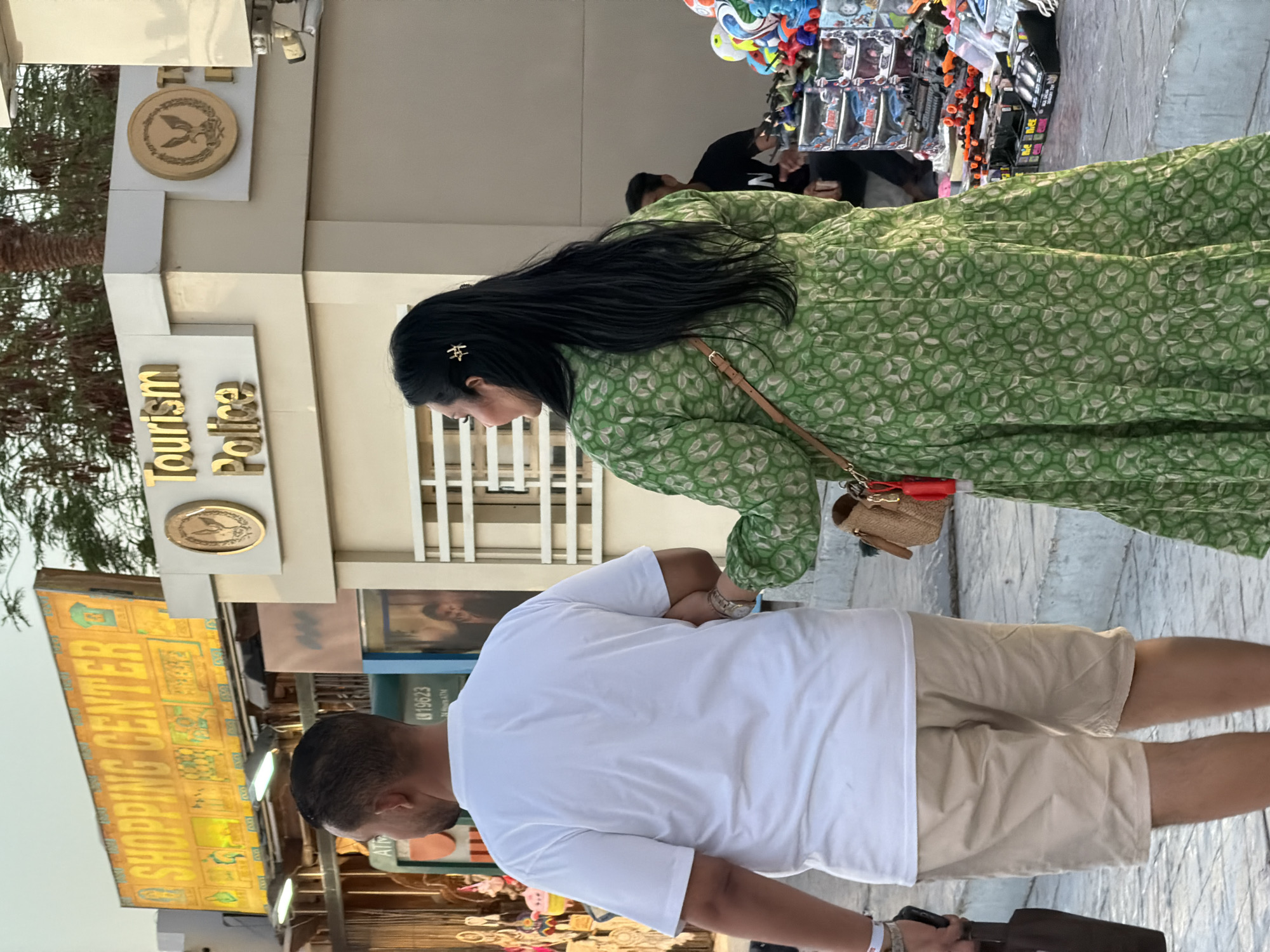 In Sharm El Sheikh, Egypt, two people, a man and a woman, are walking down a street. The man, seen from behind, wears a white t-shirt and beige shorts. The woman, also viewed from behind, is in a green, patterned maxi dress. She has long, dark hair adorned with a gold barrette. A tan crossbody bag with a red tassel hangs from her shoulder. They appear to be walking past a market stall filled with toys and souvenirs, and a building with signs for "SHOPPING CENTER" and "Tourism Police." An ATM machine is also visible. The scene suggests a typical day in a tourist area.