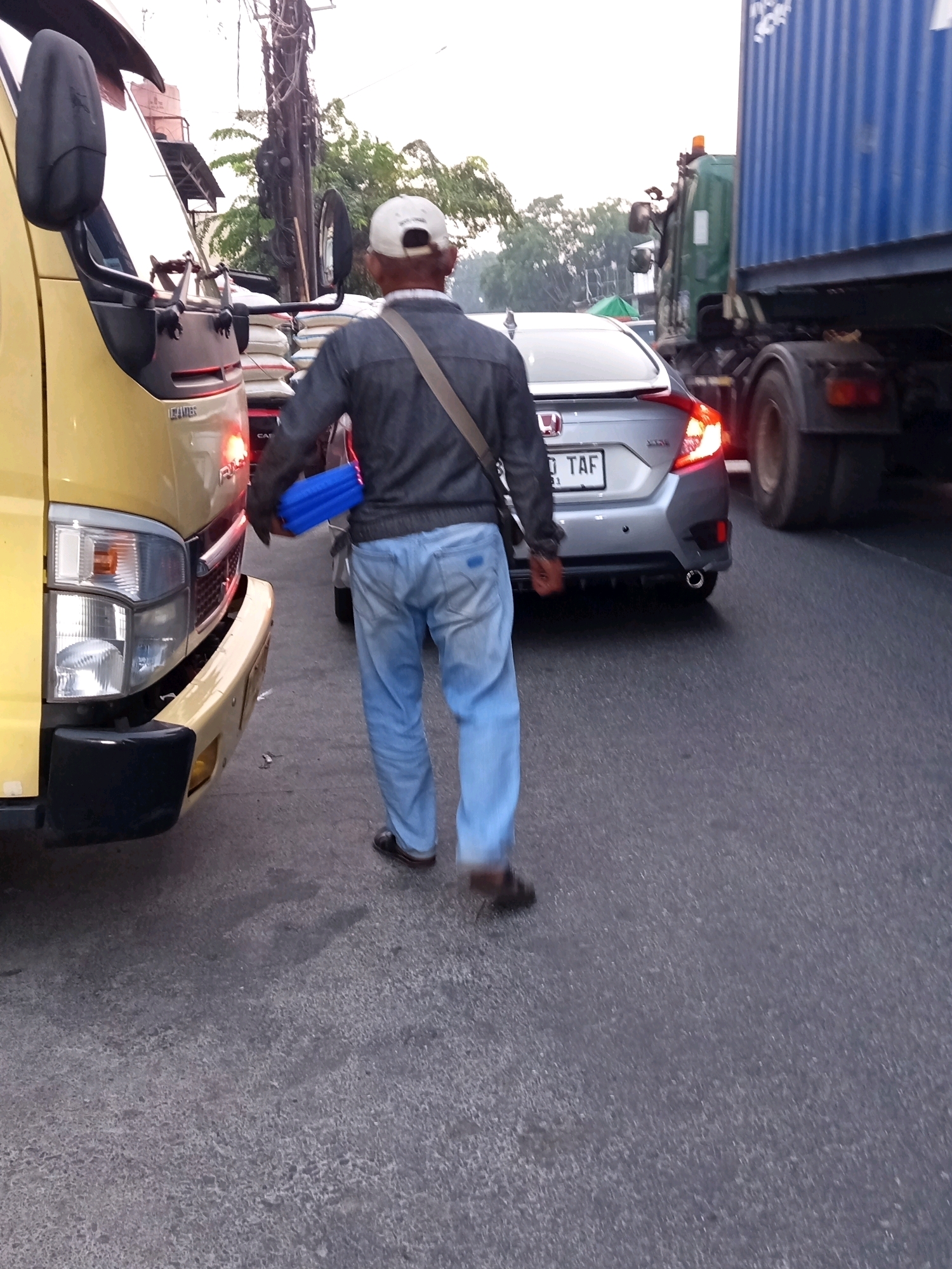 A person wearing a white baseball cap, a dark jacket, and light blue jeans walks across a street in what appears to be a traffic jam, likely in Karawang, Indonesia. The individual is carrying a bright blue cylindrical object under their right arm. In the foreground, the front of a yellow truck is visible, with its headlights and bumper showing. To the right of the person, a silver sedan with its taillights illuminated is stopped, and further to the right, a large blue shipping container truck is also stopped in traffic. The road surface is asphalt, and the sky is overcast. The scene suggests a typical urban traffic condition, possibly during daylight hours.
