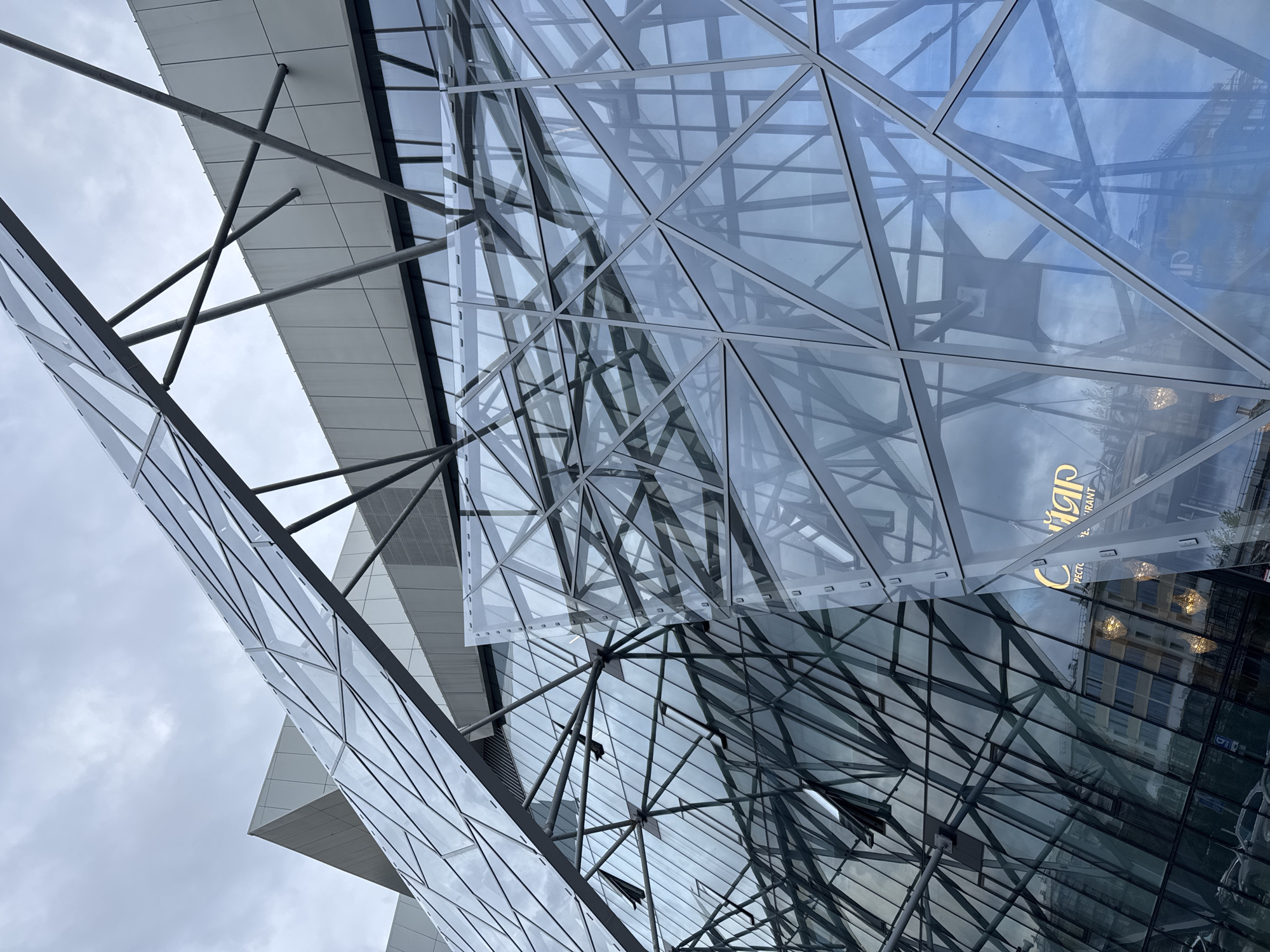 The image depicts a modern architectural structure with a geometric glass facade, likely located in Kazan, Russia. The building features a complex arrangement of triangular glass panels supported by a network of metal beams. The facade reflects the surrounding environment, including a glimpse of another building, the sky, and clouds. A restaurant sign can be seen reflected in the glass. The construction materials appear to be glass, metal, and concrete. The weather seems overcast. There are no people visible in the image.