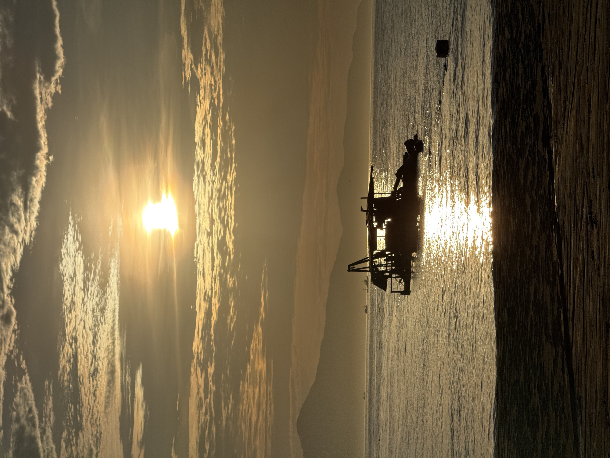 The image displays a coastal scene in Đà Nẵng, Vietnam, illuminated by the low-angle sun, suggesting either sunrise or sunset. The sky, predominantly golden and hazy, features a bright, overexposed sun in the upper-left quadrant, with scattered altocumulus clouds. A range of dark, silhouetted mountains stretches across the horizon, partially obscured by atmospheric haze. The calm ocean surface reflects the sun's golden light, creating a shimmering path extending towards the viewer. In the mid-foreground, a dark, silhouetted boat, possibly a fishing or dredging vessel with an A-frame structure on its deck, rests on the water. Several smaller, distant boat silhouettes are visible near the mountain base. To the right of the main boat, a dark, cylindrical object floats on the water. The immediate foreground consists of a dark, textured sandy beach or shoreline, exhibiting subtle ripples where it meets the water.