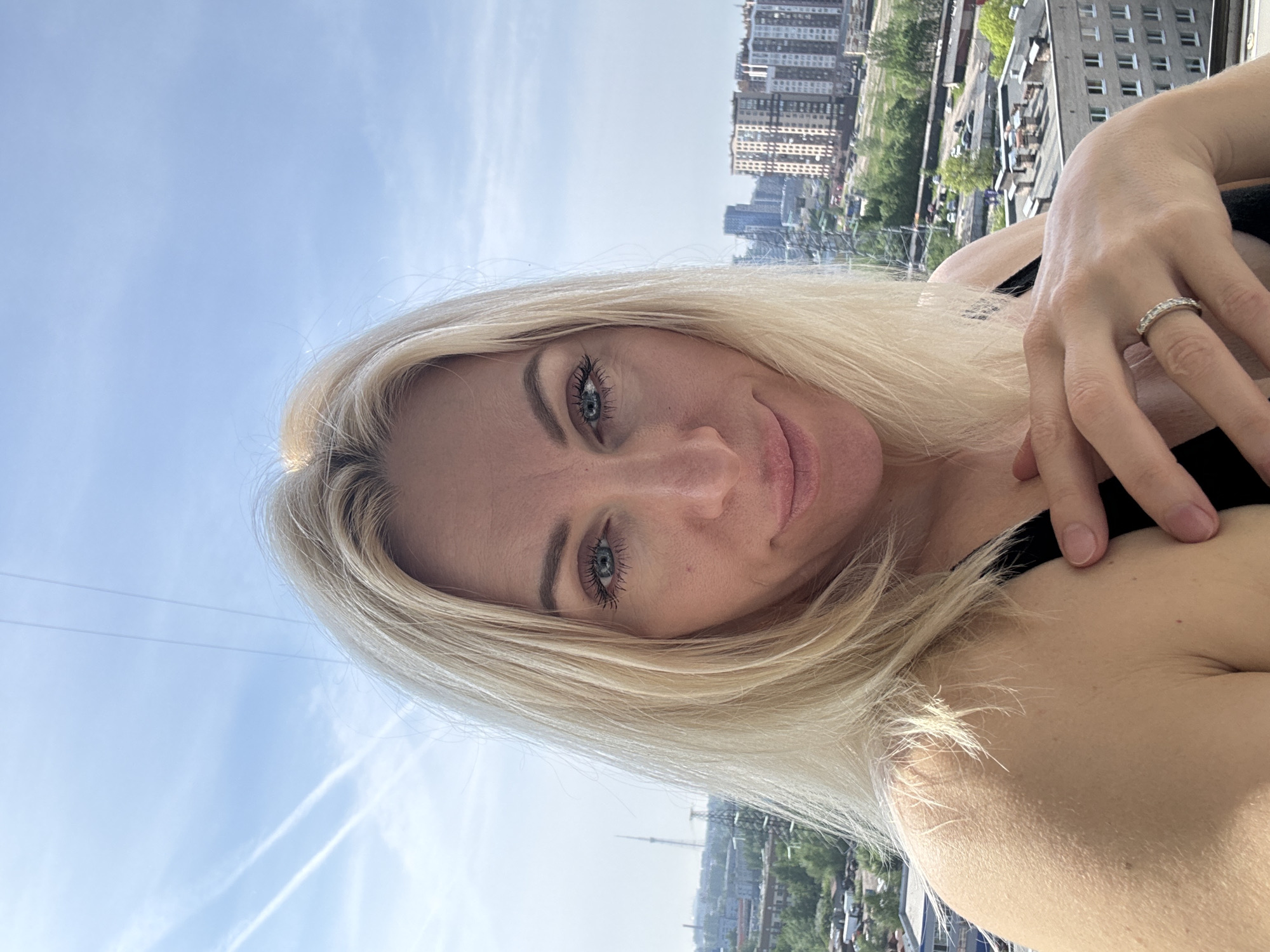 A woman with blonde hair and blue eyes takes a selfie. She is wearing a ring on her left ring finger. The background includes buildings and trees. The sky is blue with some clouds. The city is Unknown, Unknown.