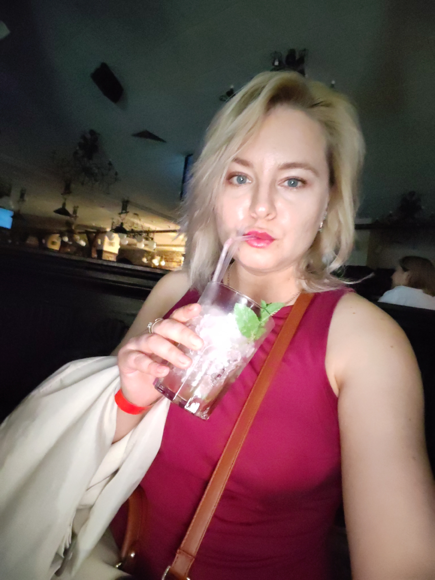 A Caucasian woman with blonde hair, wearing a pink dress and a light-colored shawl, is taking a selfie while holding a glass with a pink beverage and a straw. She is wearing a red wristband and a ring on her left hand. She is in an indoor setting, likely a bar or restaurant, with a dimly lit ceiling and background decor. The woman's face is turned towards the camera, and her lips are slightly parted as she sips the drink. In the background, another person with brown hair is visible, seated at a table. The environment suggests a casual evening outing.