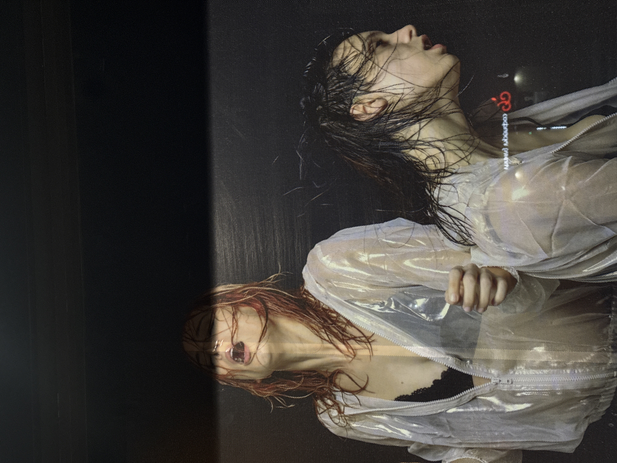 The image shows two side-by-side portraits of women with wet hair. The woman on the top has dark, wet hair. The woman on the bottom has reddish-brown, wet hair. Both women are wearing a translucent zippered jacket, unzipped to reveal a black garment underneath. The background is dark. The words "MARIA MAXIMOFFA CONCEPT GALLERY" appear in red neon writing on the right side of the upper portrait. The overall aesthetic suggests a fashion or art context. Based on the text, the location is Sofia, Bulgaria.