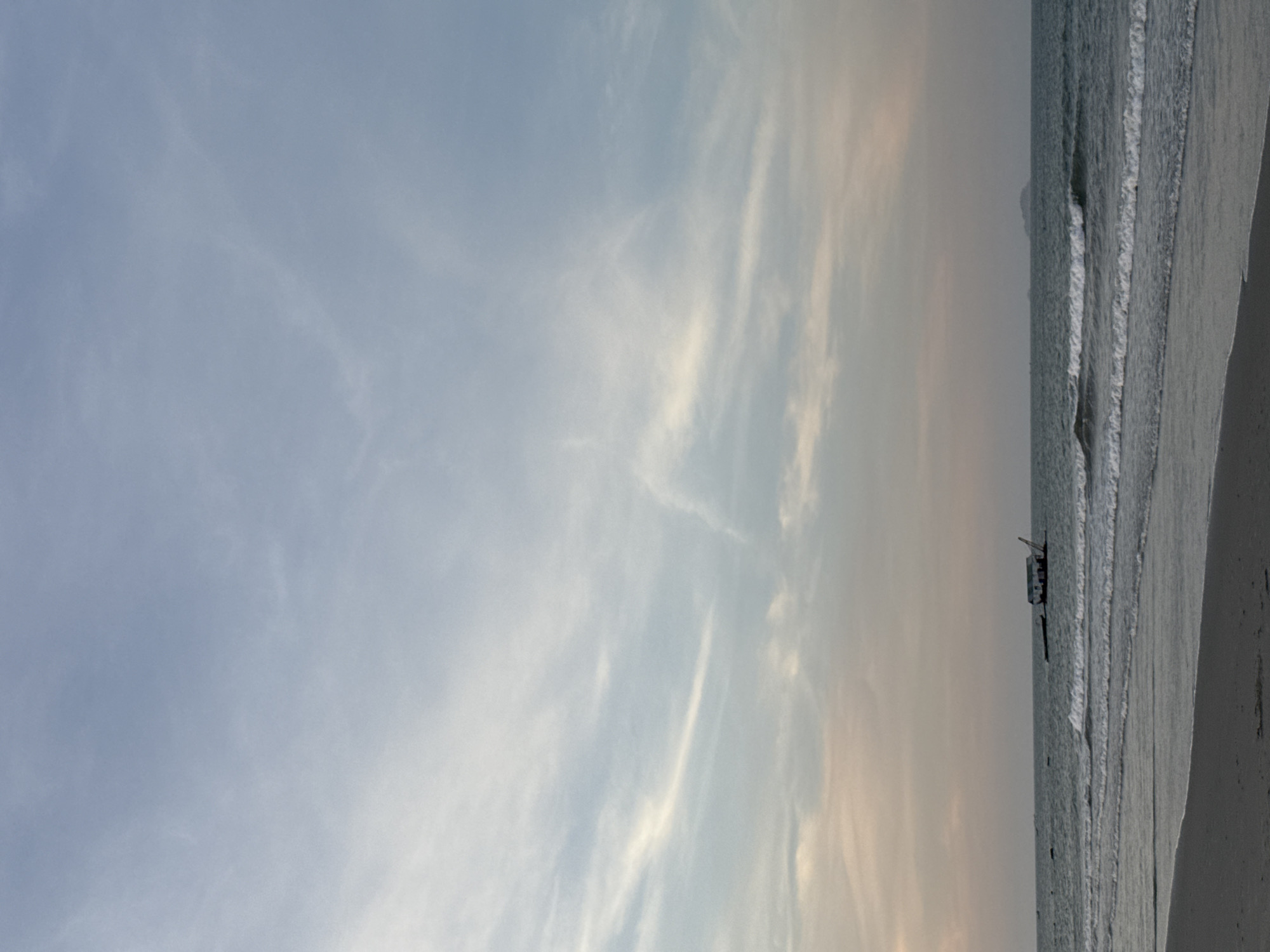 The image shows a beach scene in Đà Nẵng, Vietnam. The sky is partly cloudy with a mix of blue and light orange hues. The ocean meets the shore with gentle waves. A small boat or structure is visible in the water close to the shoreline. The sand is pale and slightly wet. No people are visible in the frame.
