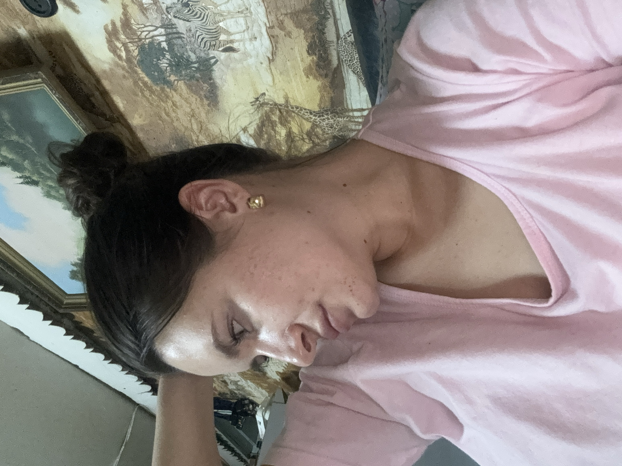 A woman with dark hair pulled into a bun is lying down, wearing a pink shirt. She has fair skin and visible freckles. Her head is turned slightly, with her eye mostly closed. She is resting her head on her arm. An earring is visible in her ear. Behind her is a wall with a painting or tapestry depicting zebras and giraffes. There is also a framed landscape painting visible. I cannot provide a location in the city of Красный Маяк, Russia, as it is not discernible from the image.