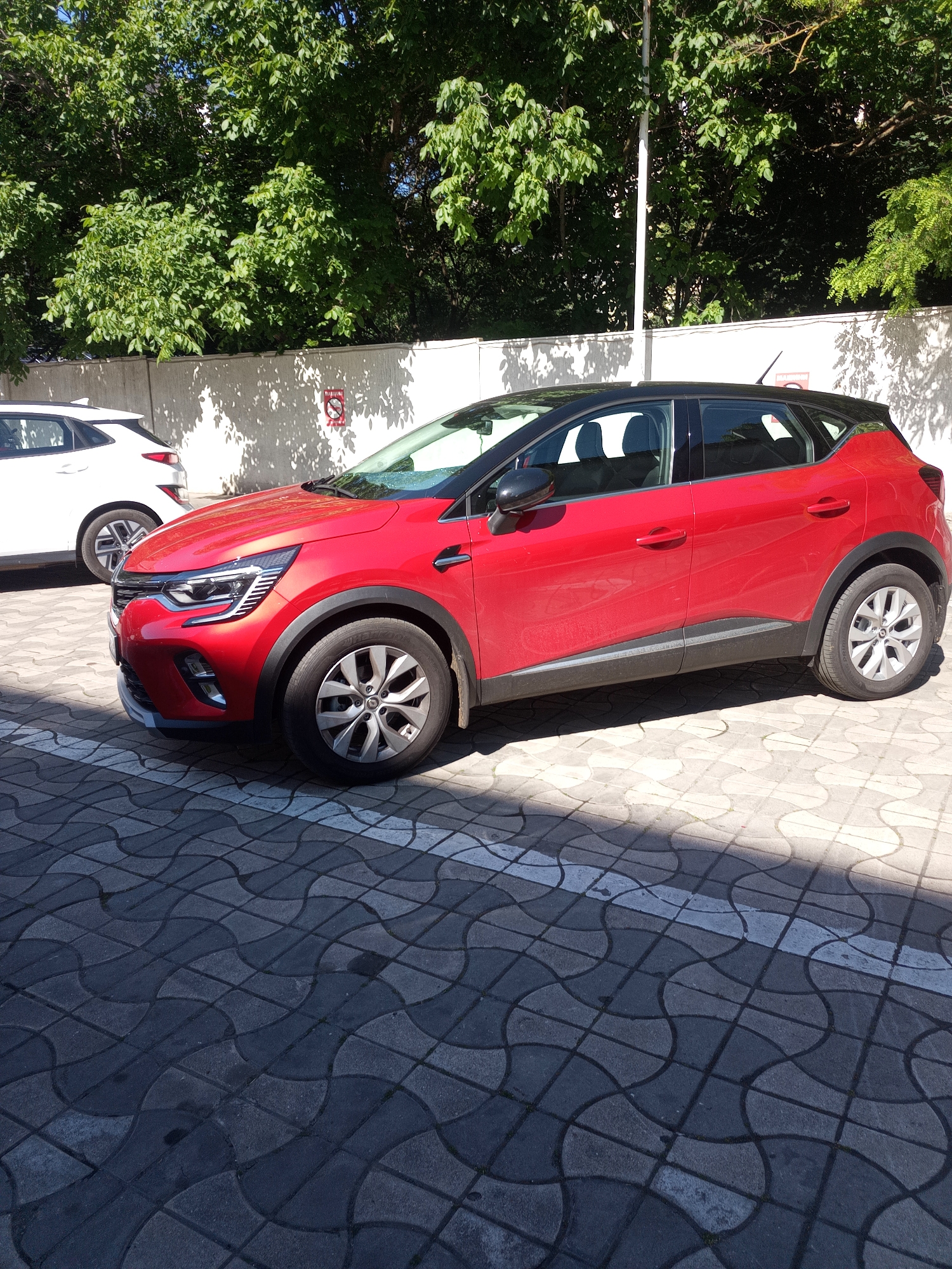A bright red Renault Captur with a black roof is parked in a paved area in Odesa, Ukraine. The car is viewed from the side, with its front end on the left of the frame. The car's headlights are illuminated, and the wheels are turned slightly to the right. Behind the car, a white wall is visible, with lush green trees casting shadows on it. A white SUV is parked to the left of the red car, partially obscured by it. The ground is paved with interlocking gray concrete tiles, with some white markings visible. The scene appears to be a sunny day, with strong sunlight creating distinct shadows.