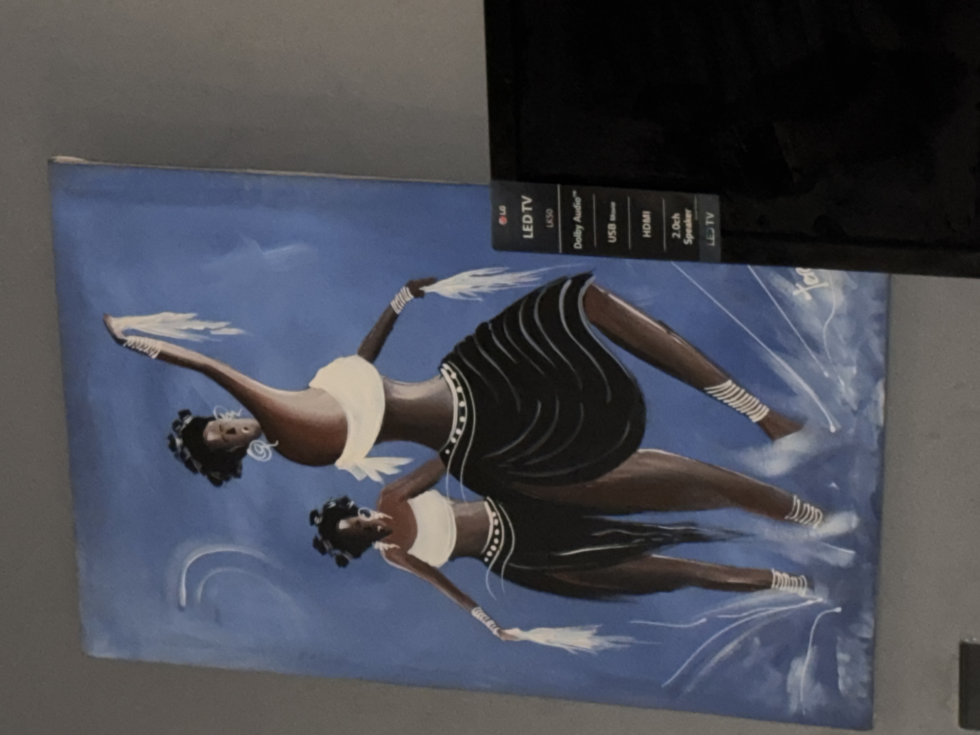 The image shows a painting depicting two dark-skinned women in traditional attire. The women are positioned in a dynamic pose, seemingly dancing or in motion, against a blue background. They wear white tops and black skirts adorned with white accents. Both figures have white decorations on their arms and legs, and hold white feathered fans. The background is predominantly blue with white strokes suggesting movement or a dynamic environment. The painting is positioned near an LG LED TV, partially obscuring its details, but the TV's ports (USB, HDMI) are visible. The context of the painting is not specifically Kurunduma, Nigeria.