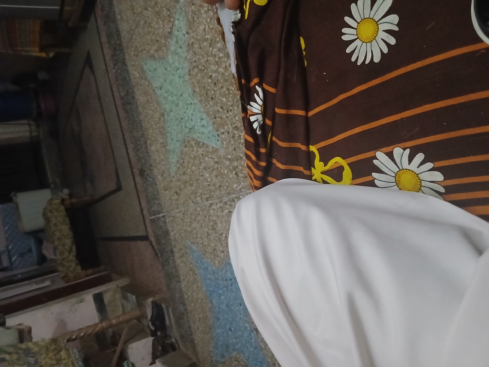 The image depicts an indoor scene, likely a living area, with a focus on the ground and a person's draped leg. The floor is a patterned terrazzo with inlaid star shapes, one blue and one light green. A portion of a person's leg, covered in a white fabric, occupies the left side of the frame. To the right, a patterned blanket or fabric with a brown background, orange stripes, and white daisy-like flowers is visible. In the background, elements of a room are blurred and out of focus, including what appears to be furniture, a wooden structure resembling a chair or table leg, and a patterned rug. The lighting is dim, suggesting an evening or indoor setting. The scene is static and does not show any active interactions. The provided context suggests this location is in Peshawar City Tehsil, Pakistan.