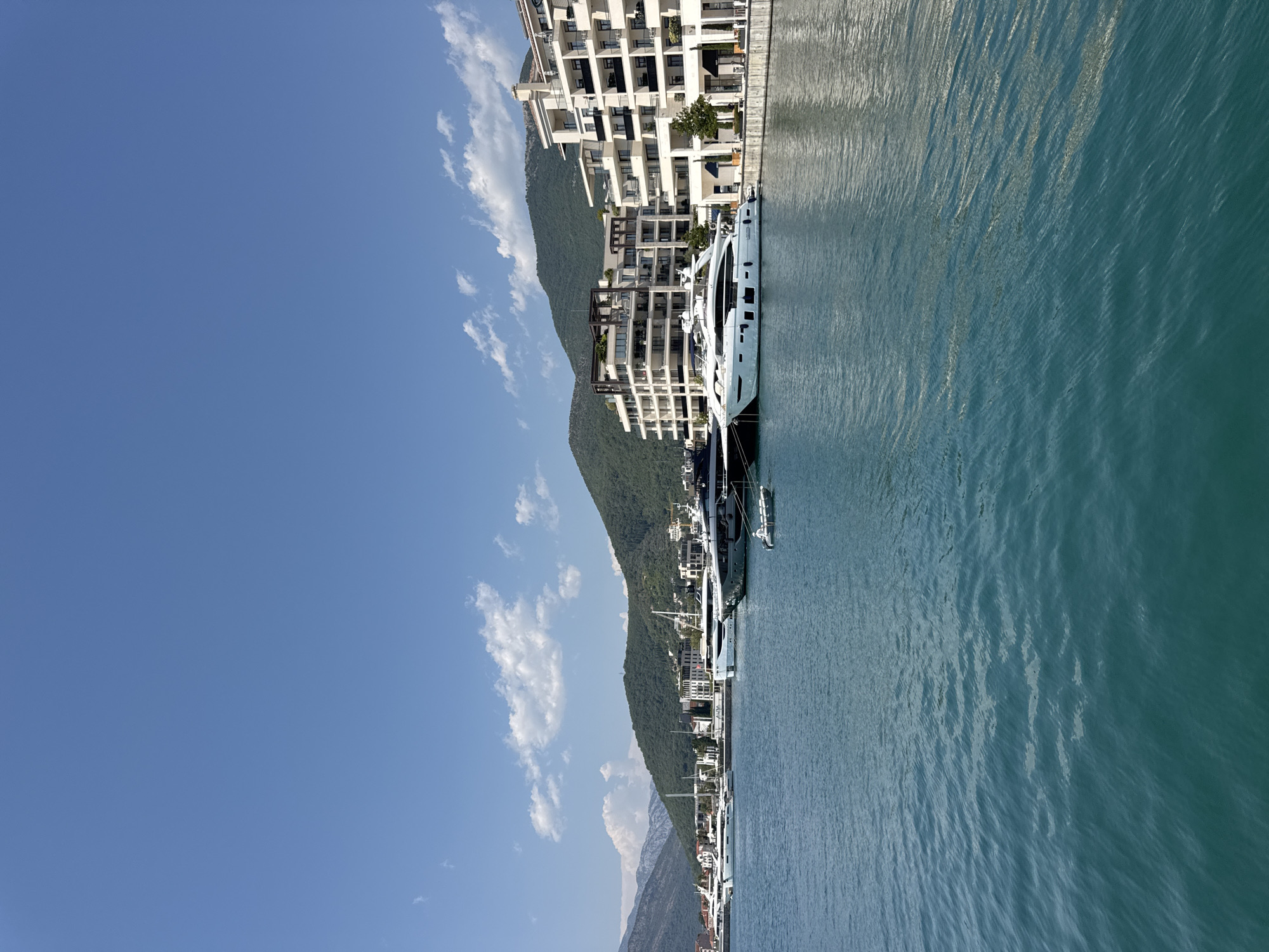 The image shows a marina in Tivat, Montenegro, with several yachts docked along the waterfront. The water is a deep turquoise color and appears calm with gentle ripples. Behind the yachts, there are modern white buildings with multiple balconies, suggesting a developed resort area. Green, tree-covered mountains form the backdrop, rising steeply from the shoreline. The sky is a clear, bright blue with a few scattered white clouds. The scene suggests a sunny day in a picturesque coastal city.